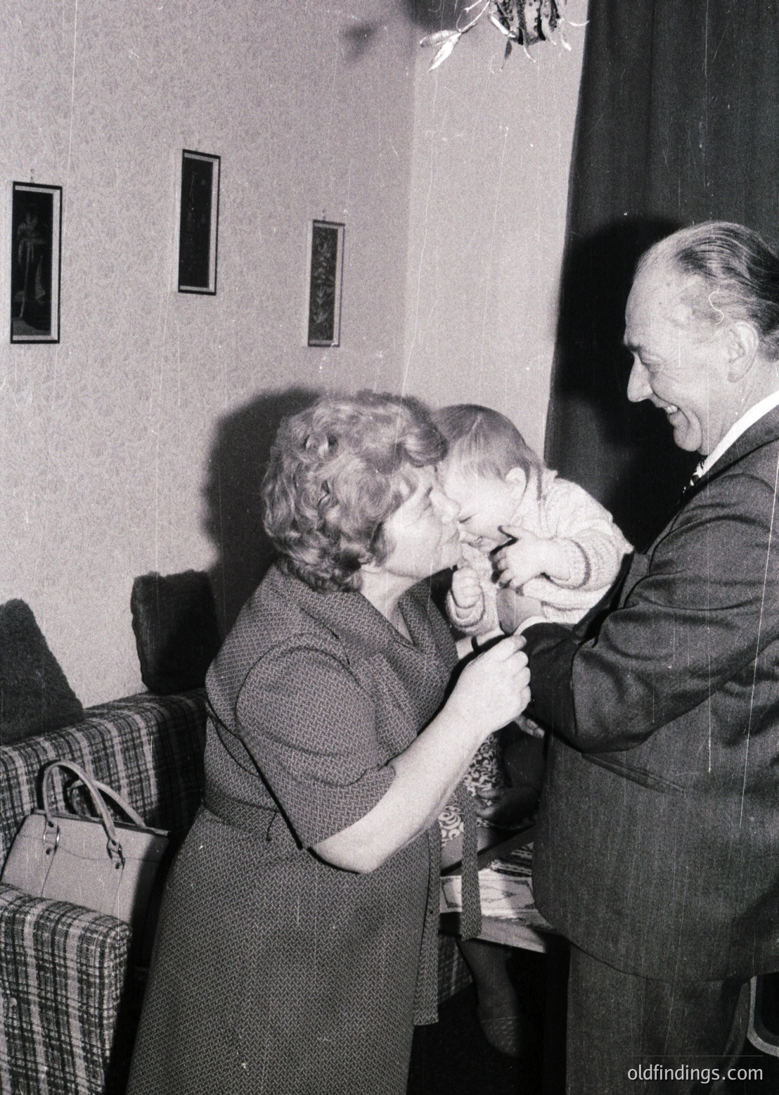 A woman in a dark dress tenderly kisses a child’s forehead while an older man in a suit smiles, holding the child’s hand. Indoor setting with patterned wallpaper and a checkered armchair in background. Likely mid-20th century (1950s–1960s) based on clothing and decor.