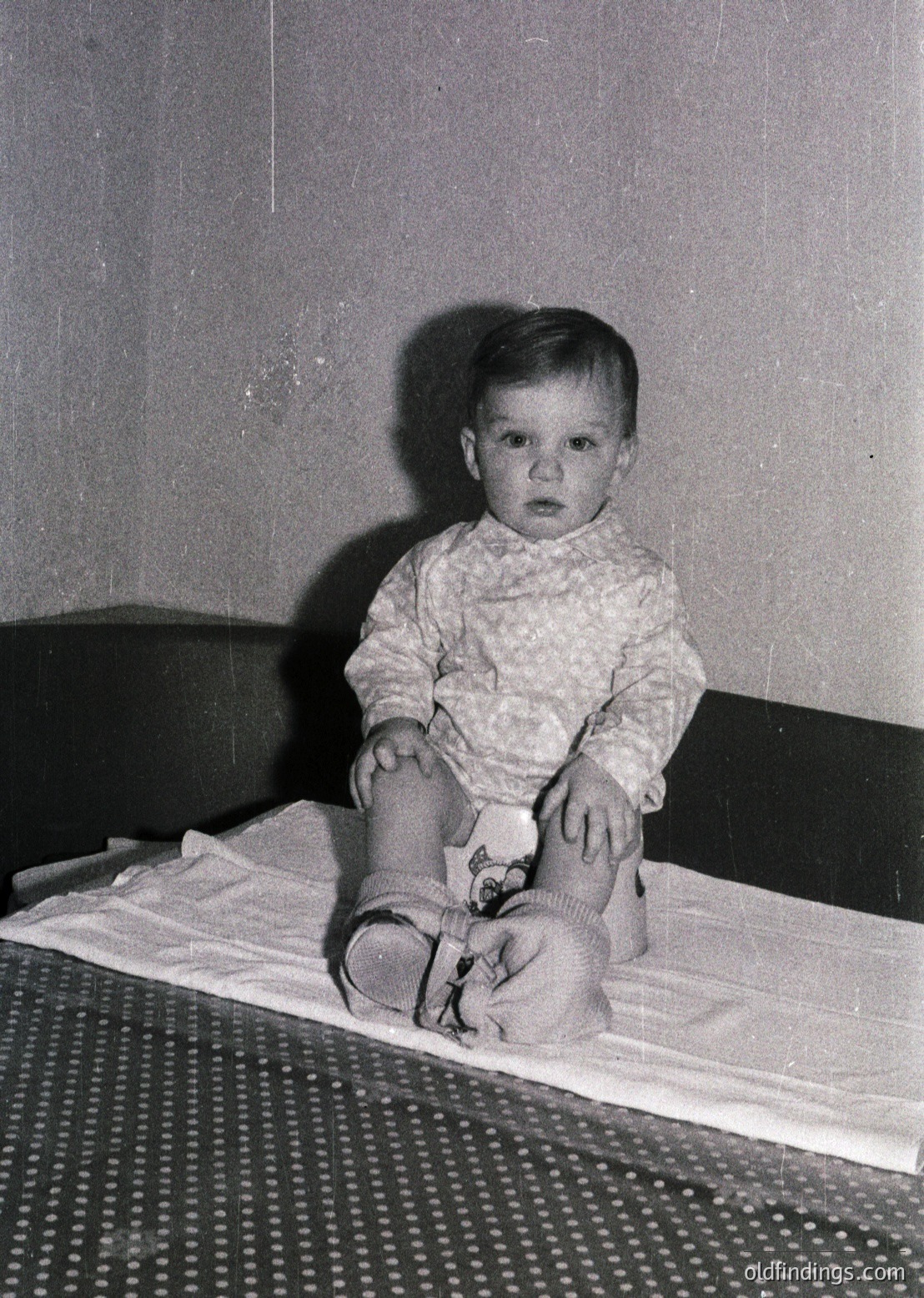 Young child in mid-20th-century attire, seated on a patterned metal-framed cot with a white sheet. Knitted sweater, knee-high socks, and a diaper visible. Plain interior suggests domestic setting, likely 1950s–1960s.