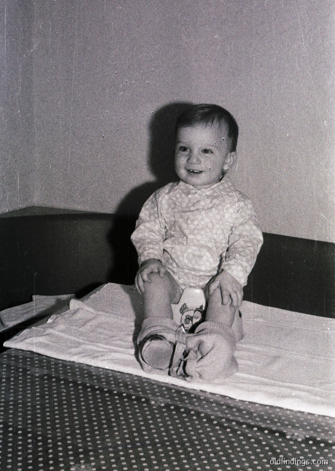 Vintage black-and-white portrait of a toddler sitting on a patterned blanket, wearing a textured sweater and striped socks with cartoon characters. Indoor setting with neutral-toned wall and patterned floor. Likely mid-20th century (1950s–1960s).