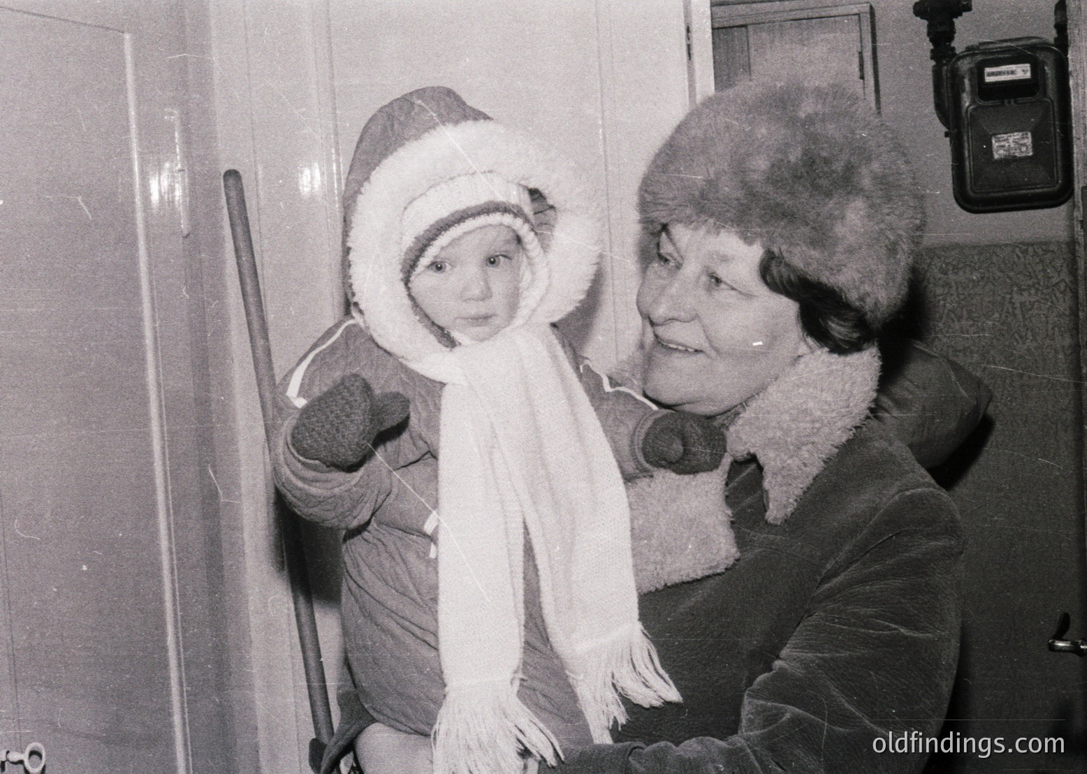 A woman in a fur-lined coat and hat holds a toddler bundled in winter attire, posing indoors near a doorframe. The child wears a knitted hat, mittens, and a scarf. The setting appears residential, likely mid-20th century.