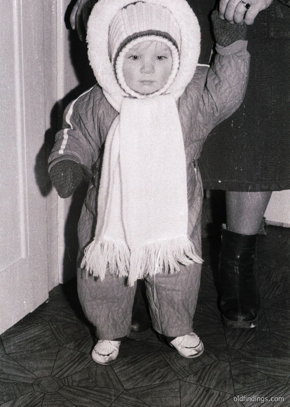 Young child in winter attire—hooded parka with fur trim, scarf, and knee-high boots—standing indoors beside an adult’s leg. Mid-20th century (1950s–1960s) fashion reflecting utilitarian winter wear.