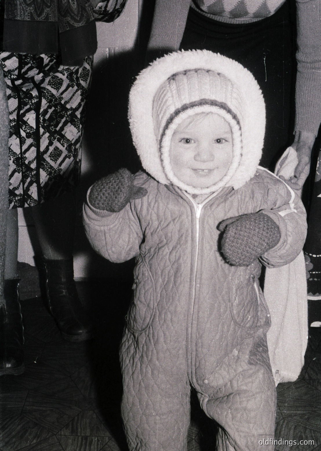 Young child in vintage winter attire, mid-20th century—puffy hooded snowsuit with mittens, posed indoors near patterned fabric. Warm, candid expression captures innocence and warmth.