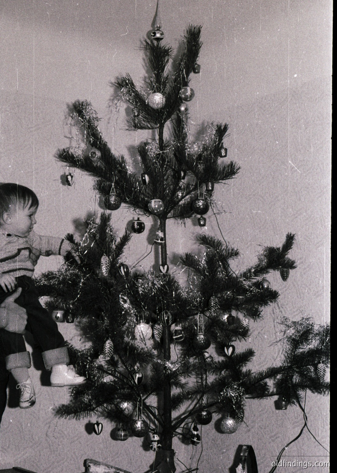 Vintage black-and-white photo of a child reaching toward a modestly decorated Christmas tree adorned with tinsel, glass baubles, and small metal bells. Indoor setting, likely mid-20th century (). Warm holiday nostalgia with simple, handcrafted decorations.