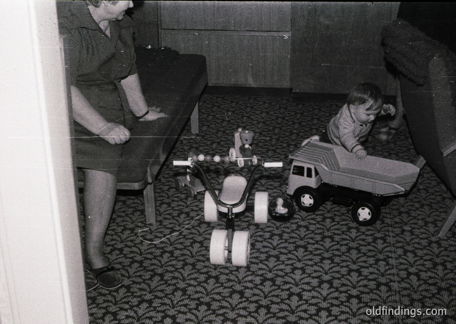 Vintage black-and-white interior shot of a woman kneeling beside a child playing with mid-century toy vehicles on patterned carpet. Motorcycle toy with large wheels and a truck toy with a child seated on it. Mid-20th century domestic setting, likely 1950s–1960s.