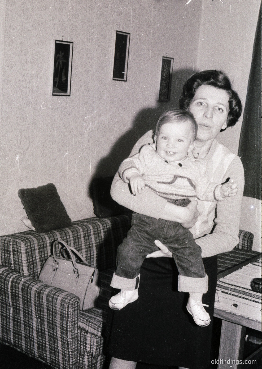 A woman cradles a toddler in a mid-century living room, likely 1950s–1960s. The child wears a striped sweater and cuffed jeans; the woman’s sweater and skirt reflect 1950s fashion. A plaid sofa, handbag, and vintage radio/record player fill the background. Warm, intimate family portrait.