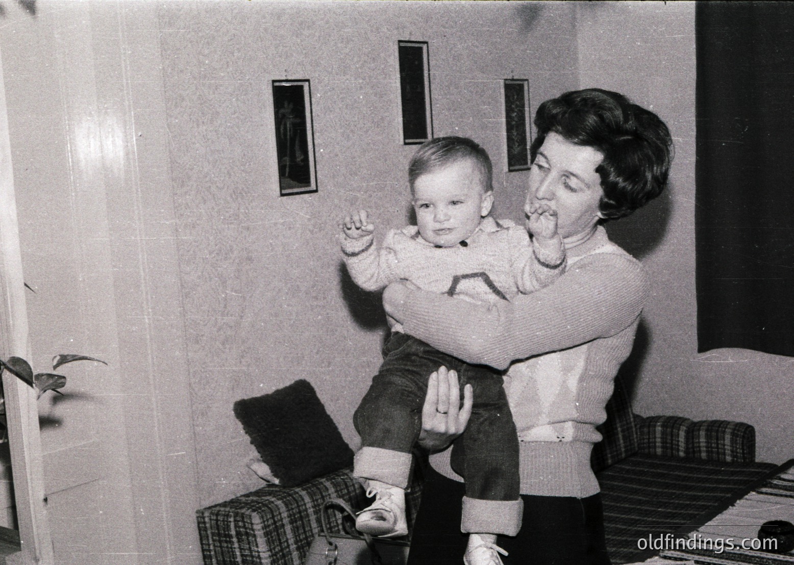 A woman in 1960s-era sweater and jeans holds a toddler on her lap in an indoor setting. Both wear layered clothing, suggesting cooler weather. The room features simple, functional decor: a checkered pillow, a potted plant, and a window with vertical blinds. The candid moment captures a tender, everyday interaction.