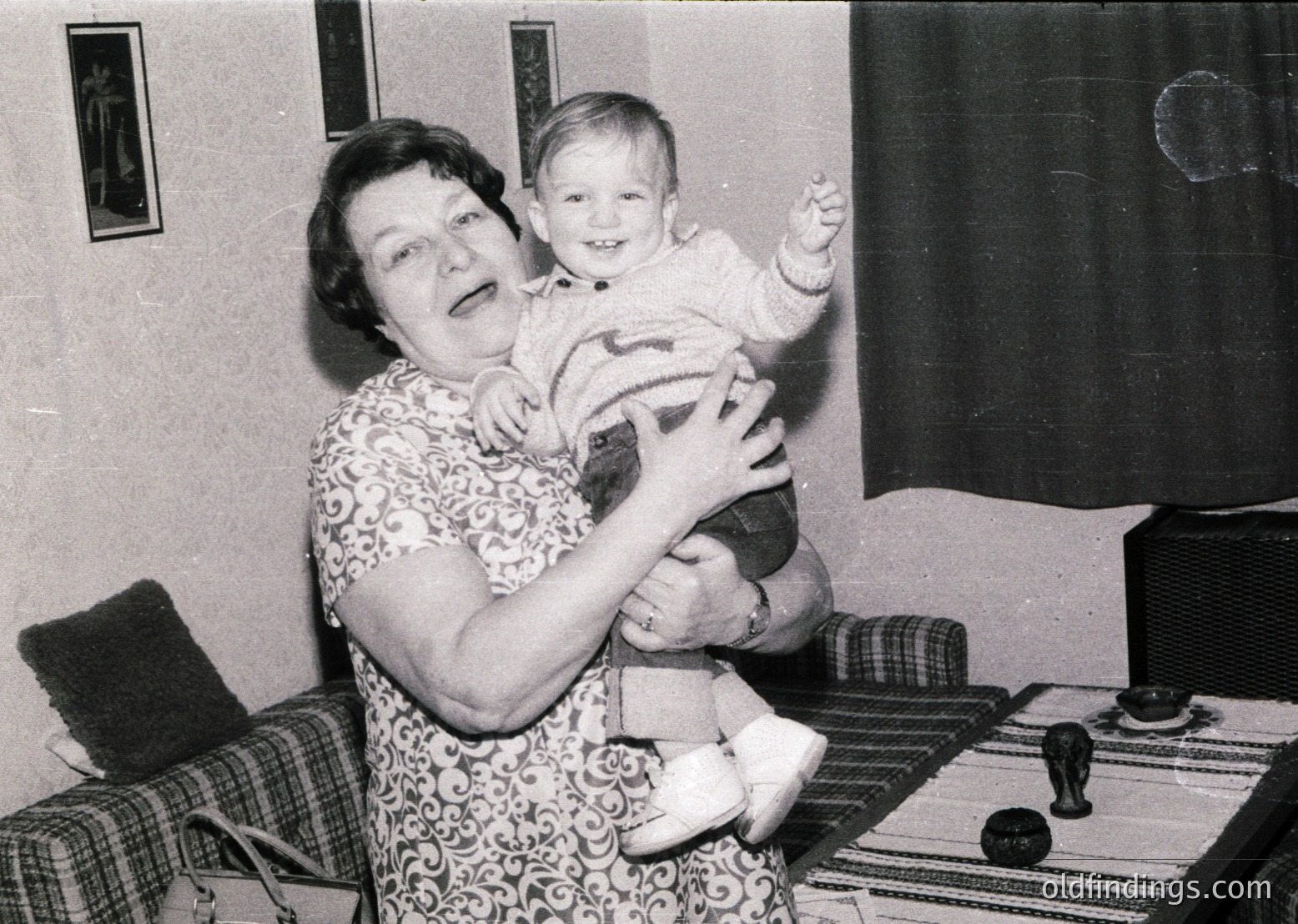 Mid-20th century indoor portrait: woman in floral-patterned dress cradles a toddler in a striped sweater, seated on a patterned rug beside a piano. Black-and-white, likely 1950s–1960s.