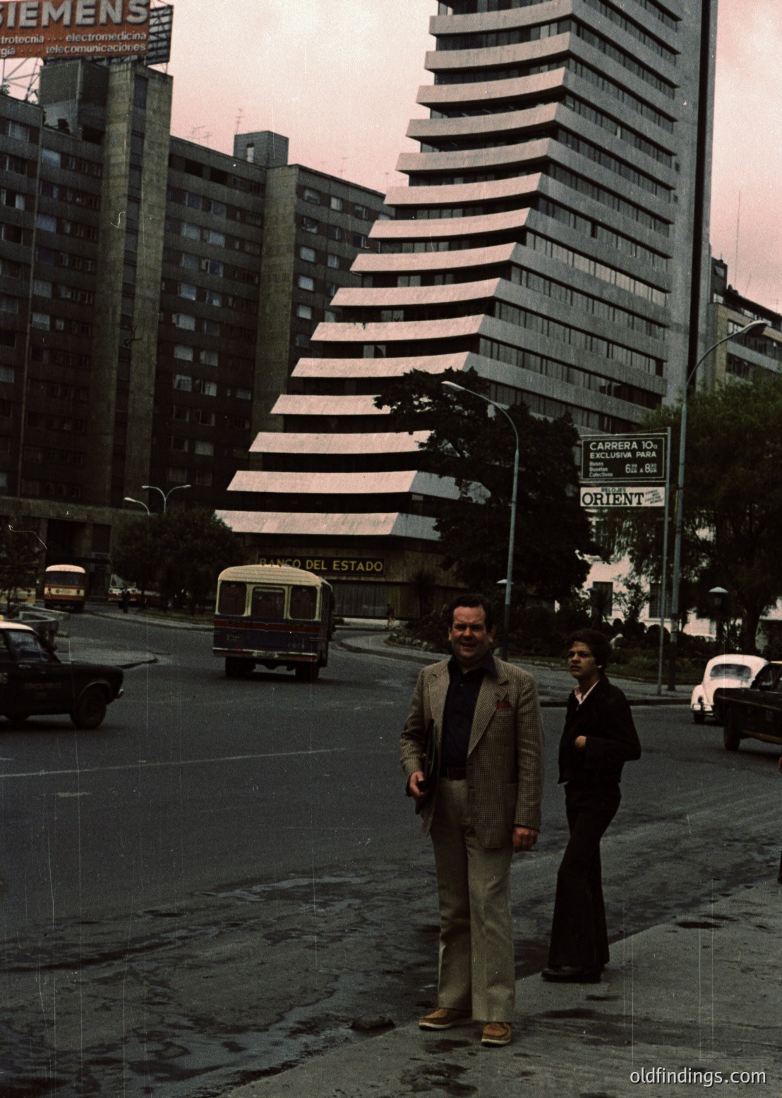 Two men in 1970s suits pose on a wide, empty urban street in Mexico City. The striking **Torre Latinoamericana** (built 1956) dominates the background, its Brutalist concrete and horizontal stripes iconic. A vintage bus and cars, including a Siemens sign, suggest mid-century infrastructure. Street signs indicate **Calle Oriente**—likely near **Paseo de la Reforma**.