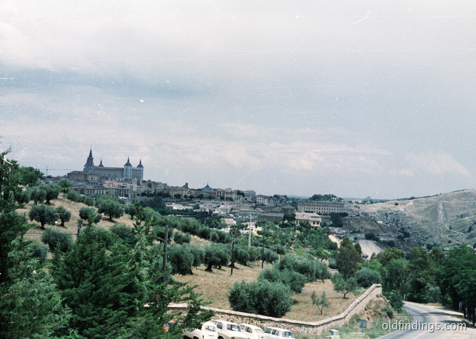 Vintage aerial view of Toledo’s historic skyline, featuring its iconic cathedral perched atop a hill. Olive groves and medieval architecture blend into the hilly terrain, showcasing Spain’s cultural heritage. Likely captured in the mid-20th century ().
