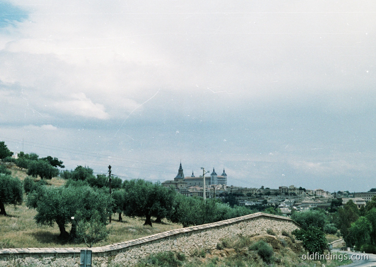 Vintage aerial view of a hillside town with a prominent Baroque-style church atop a hill, surrounded by dense olive groves. Stone retaining walls and winding roads frame the landscape. Likely