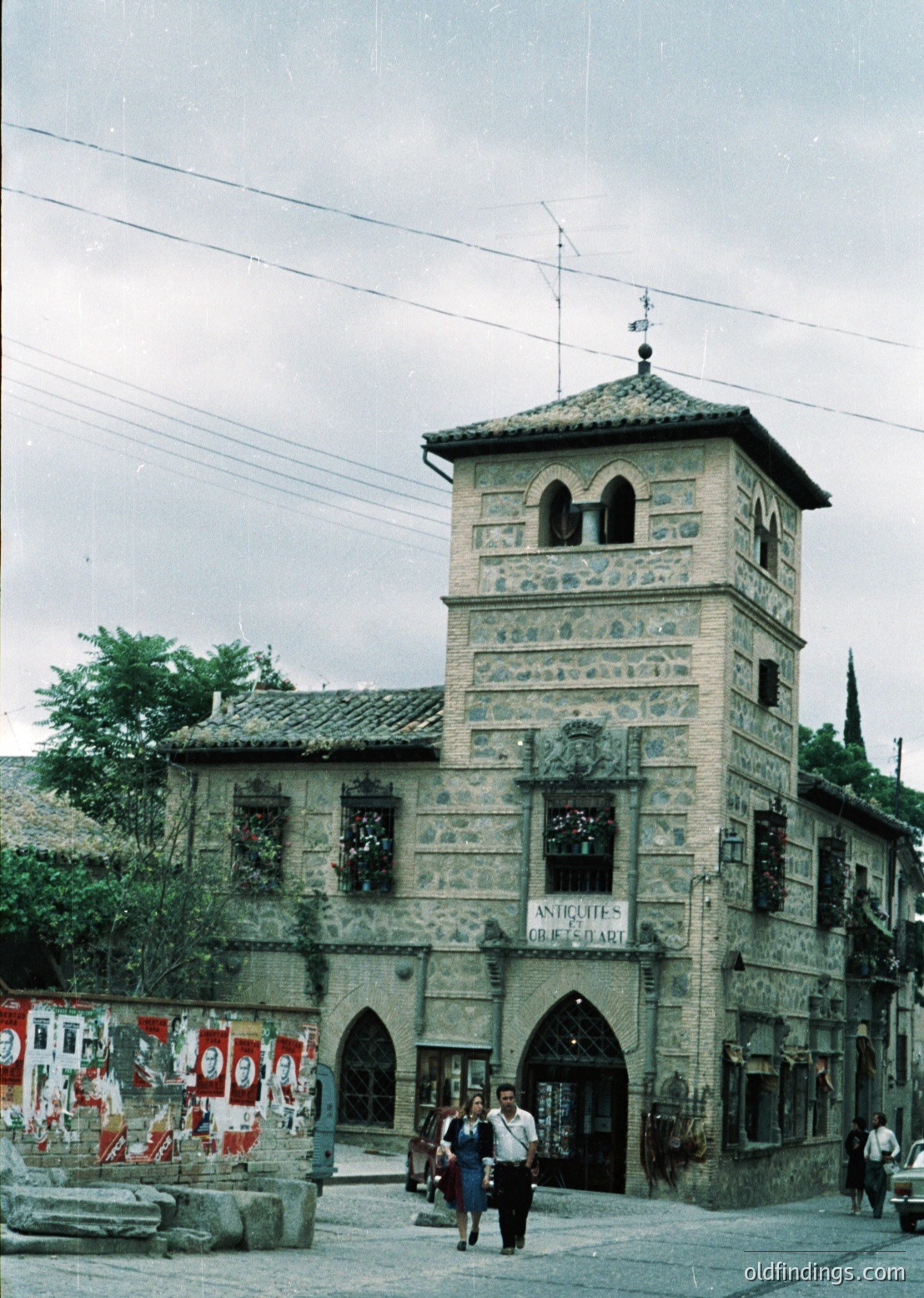 Historic stone tower with Gothic Revival details, likely a 19th-century landmark. The building features arched windows, decorative carvings, and a sign reading "Antiques & Art." Urban street scene with pedestrians and vintage signage.
