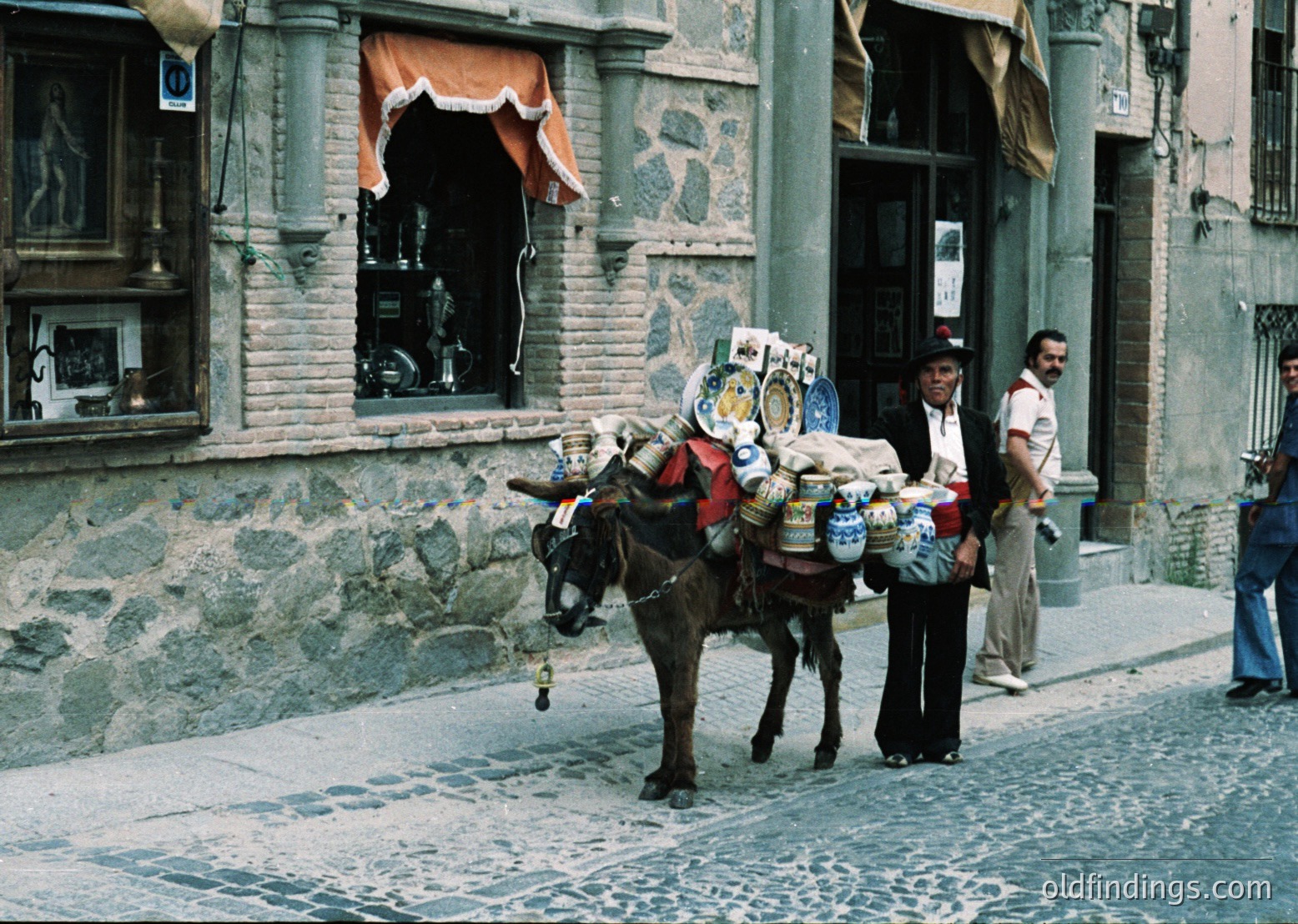 Vintage street scene featuring a donkey laden with colorful crates and sacks, likely transporting goods in a historic European town. Stone buildings with arched windows and cobblestone streets suggest 19th–early 20th century urban life. Man in traditional attire guides the donkey; pedestrians in period clothing add authenticity.