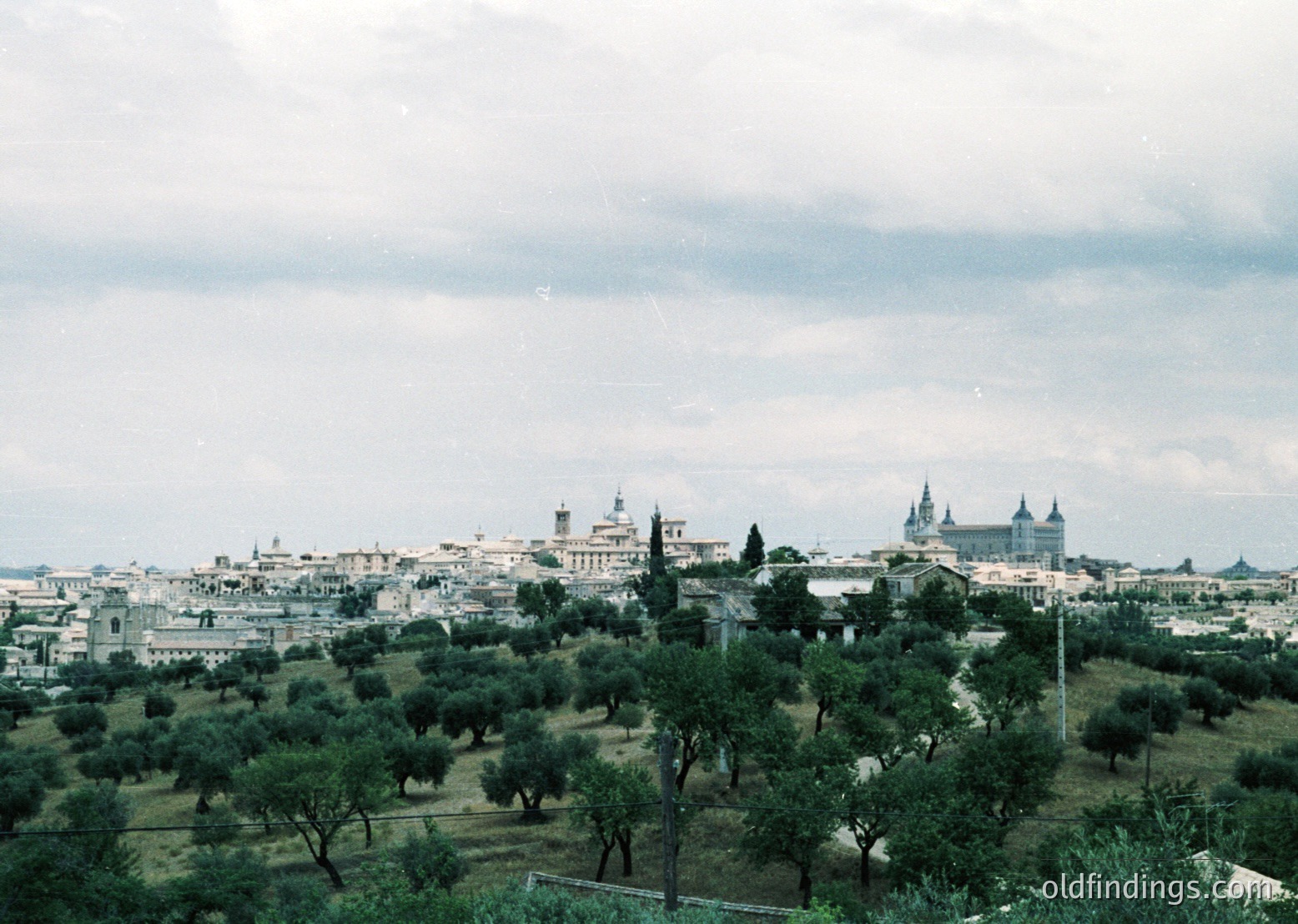 Vintage aerial view of Madrid’s skyline, featuring the Alcázar of Toledo and San Juan de los Reyes church. Lush olive groves in foreground, mid-20th century urban sprawl.