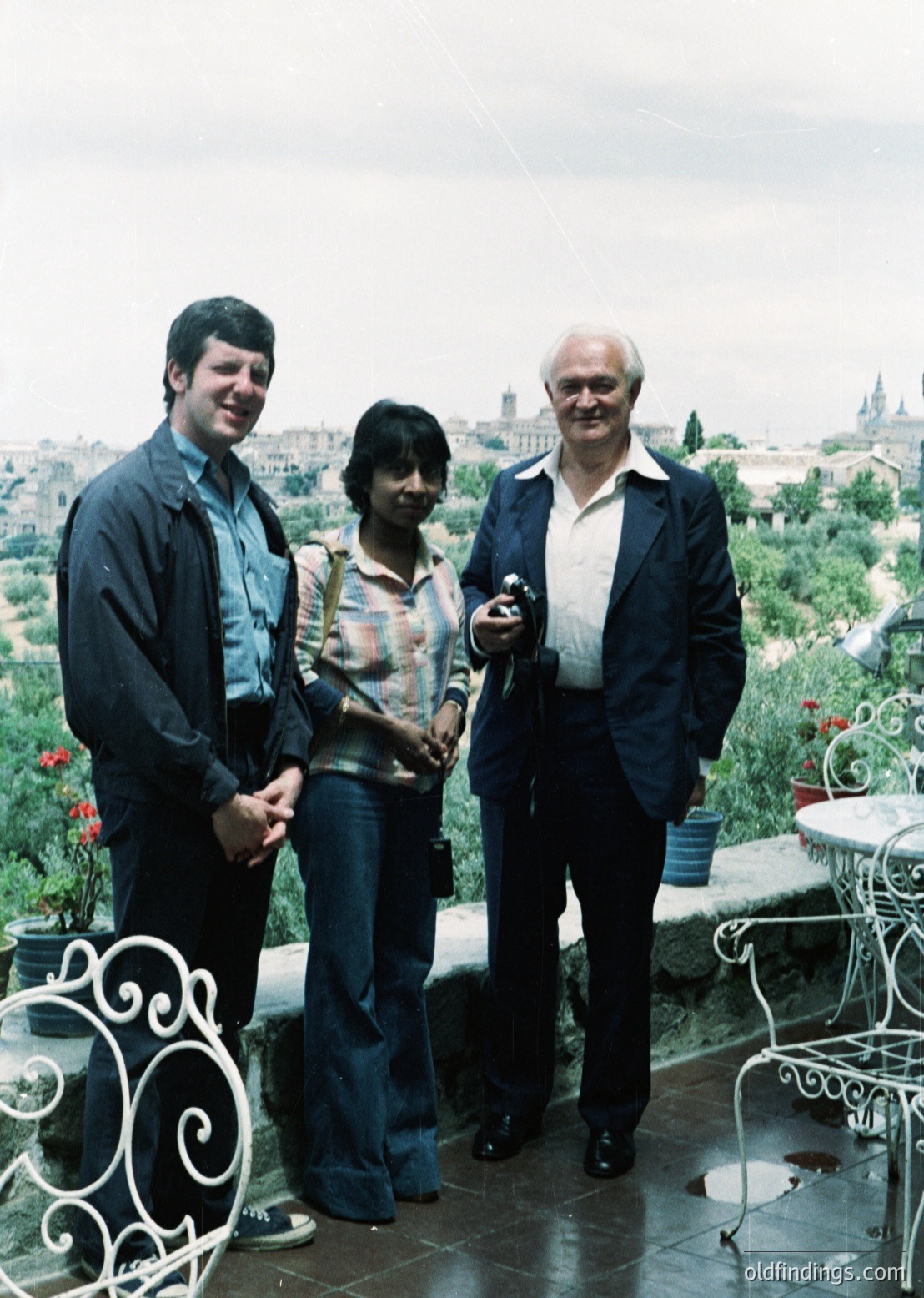 Three individuals pose on a rooftop terrace with historic cityscape in background, likely 1970s. Left: man in buttoned blazer, center: woman in patterned blouse with jeans, right: older man in dark blazer holding camera. Wrought-iron railing and potted plants visible.