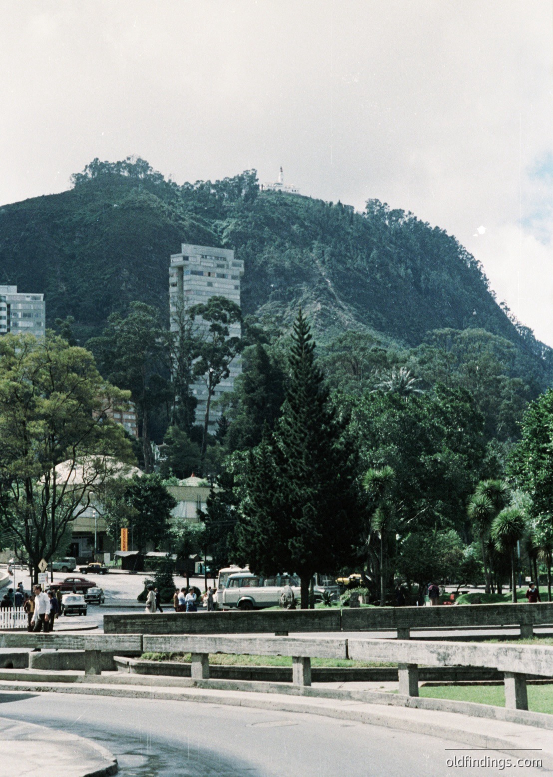 Urban park scene featuring a modernist high-rise (1960s-70s Brutalist style) perched atop a forested hill. Foreground includes a curved road, pedestrian pathways, and lush greenery. Mid-ground shows sparse vehicular traffic and a few pedestrians.