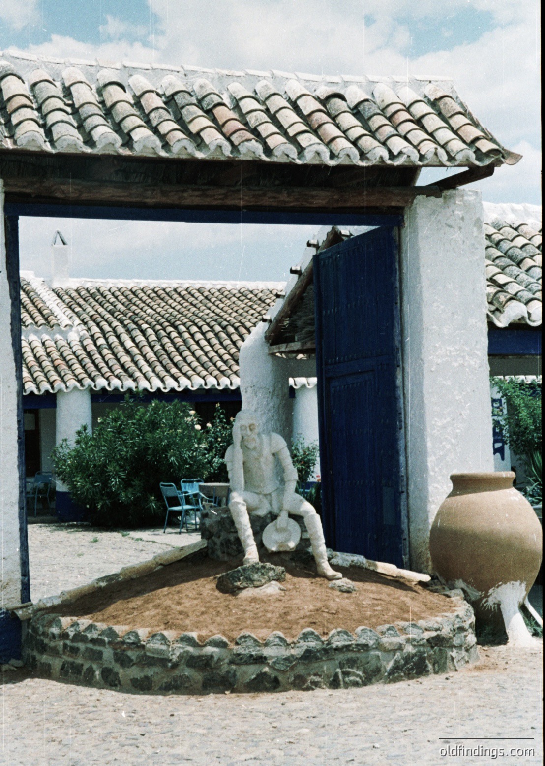 Classic Mediterranean courtyard with a sculptural fountain featuring a kneeling figure pouring water. Whitewashed walls, terracotta roof tiles, and a blue door contrast with the stone fountain base. Outdoor seating and potted plants enhance the serene ambiance.