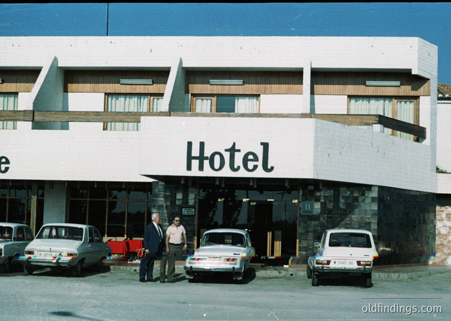 Mid-century concrete hotel façade with brutalist architectural elements—exposed stone base, angular roofline, and large "Hotel" sign in bold sans-serif letters. Two men in formal attire pose near entrance; vintage cars (1970s-era sedans) parked in front. Likely Eastern Bloc-era design, suggesting a resort or urban hotel.