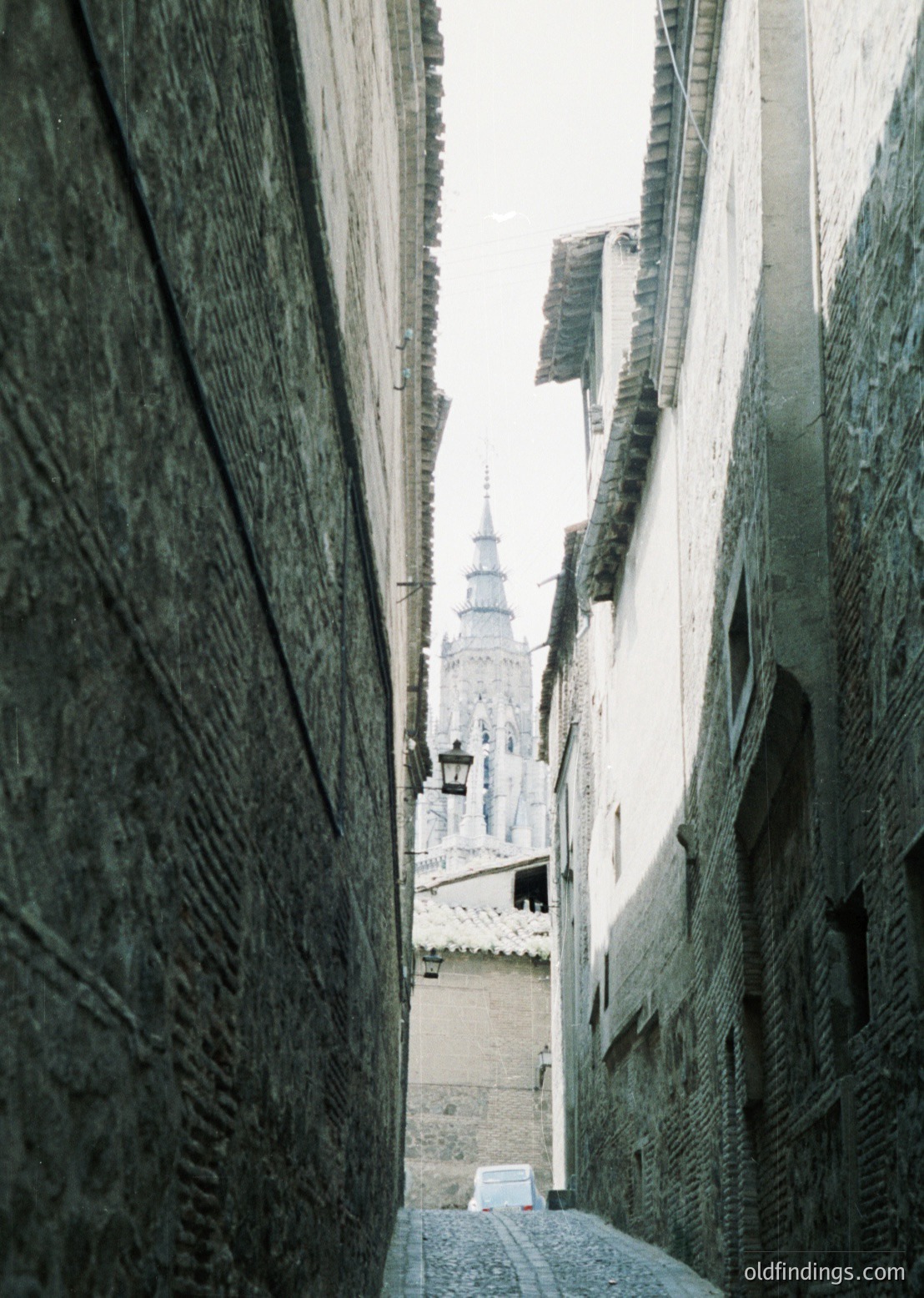 Perspective shot of a narrow, winding alley framed by aged stone walls. A historic tower with a spire rises in the distance, partially obscured by laundry hung to dry. Cobblestone path and vintage lanterns suggest a European setting, likely 19th–early 20th century.