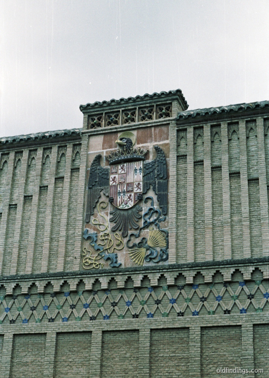 Relief sculpture featuring a crowned coat of arms with a shield displaying stacked castles and lions, flanked by seashell motifs and crowned by an eagle. Architectural facade with vertical stone columns and decorative zigzag trim. Likely Spanish colonial or neoclassical style.