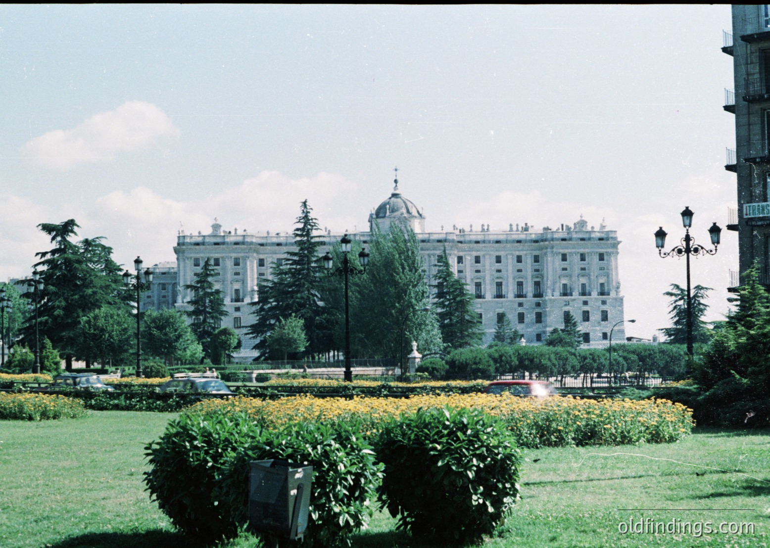 Symmetrical 1960s-era hotel with neoclassical dome, flanked by manicured hedges and flowerbeds. Classic streetlamps and vintage cars suggest mid-century urban design. Likely Eastern Bloc architecture.
