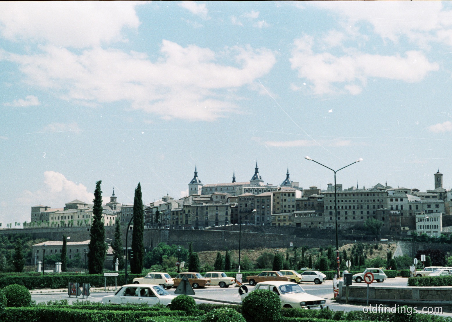 Historic Spanish cityscape featuring the Alcázar of Toledo with its iconic spires and fortified walls. Mid-20th century urban scene with vintage cars and classic streetlamps. --- *Note: The vintage cars and lighting suggest a mid-20th-century timeframe, likely the 1950s–1960s.*