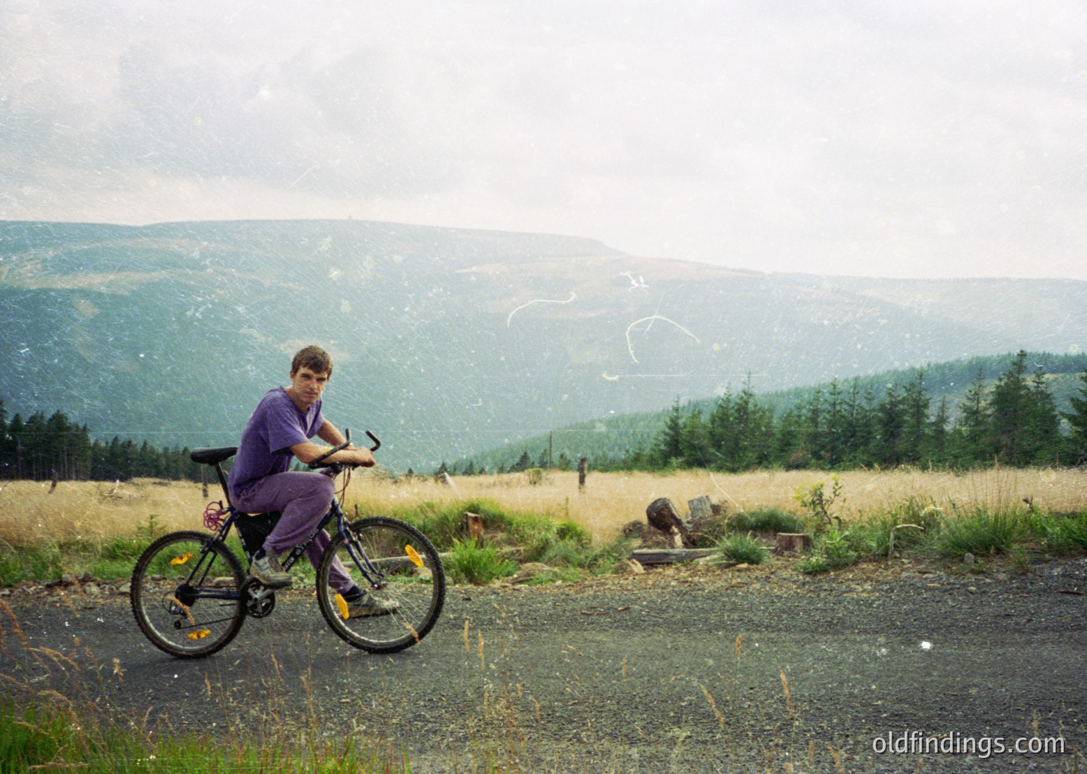 Mountain biker pauses on gravel path amid rolling hills, wearing purple long-sleeve shirt and yellow knee pads. Overcast skies and sparse forest in background suggest rural, possibly European setting. Classic 1990s mountain bike with front suspension.