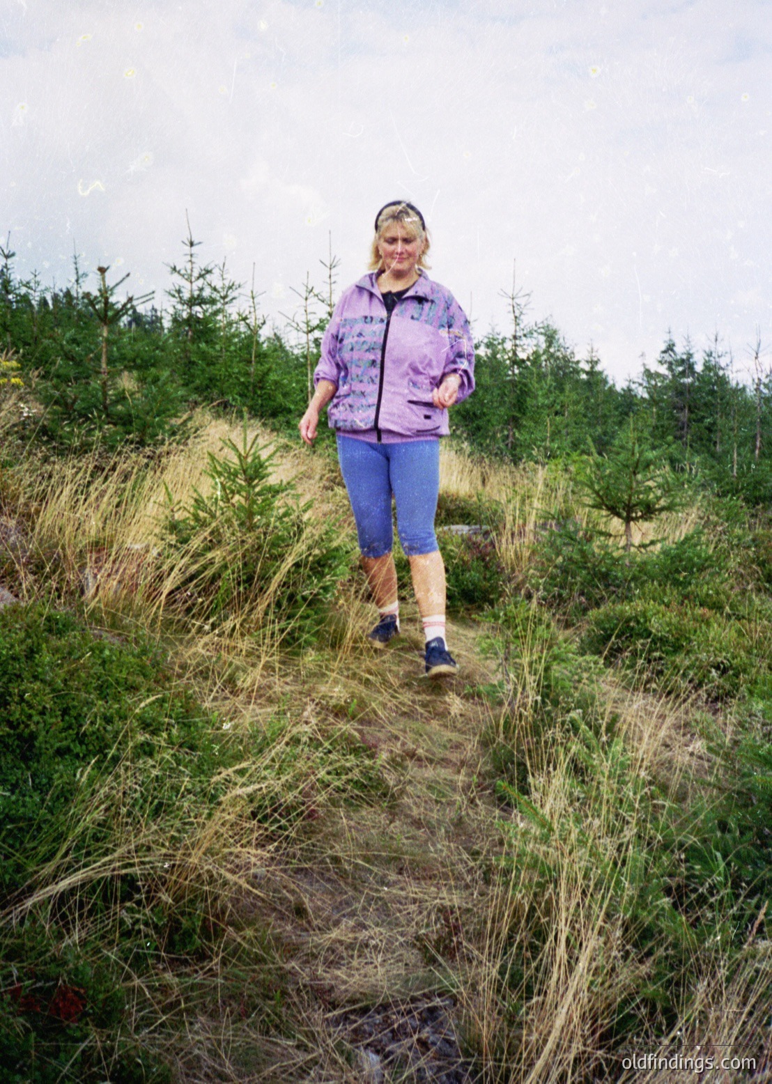 Woman in 1970s-style outdoor gear—purple jacket, high-waisted trousers, and knee-high socks—walks along a grassy, unpaved trail through a forested area. Dense greenery and pine trees frame the scene, suggesting a hike or nature exploration.
