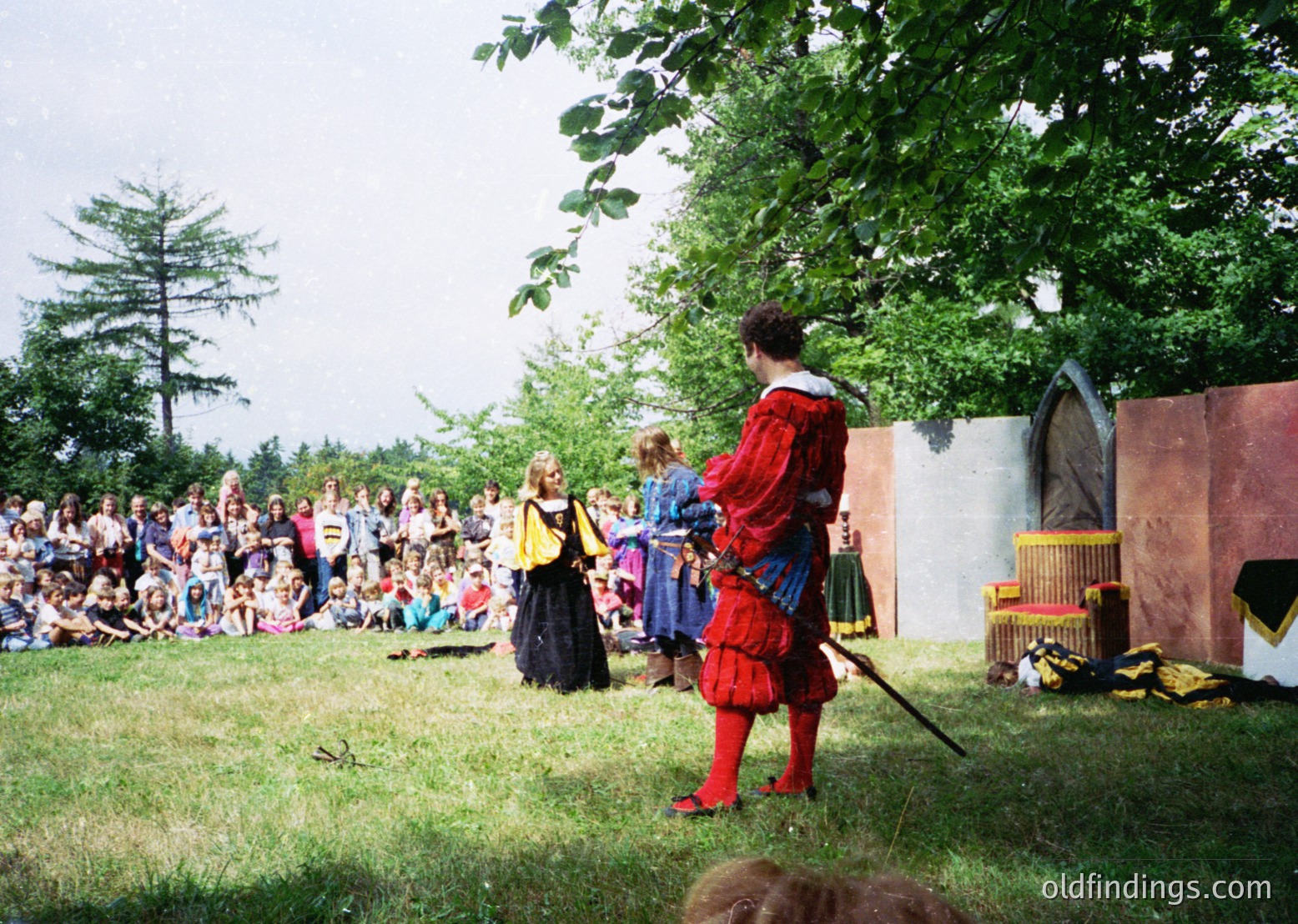 Medieval reenactment scene with participants in period attire: a figure in red tartan kilt and blue tunic holding a sword, facing a woman in dark dress with yellow sash. Crowd of onlookers in grassy outdoor setting, under trees. Likely 1990s-2000s, Western Europe.