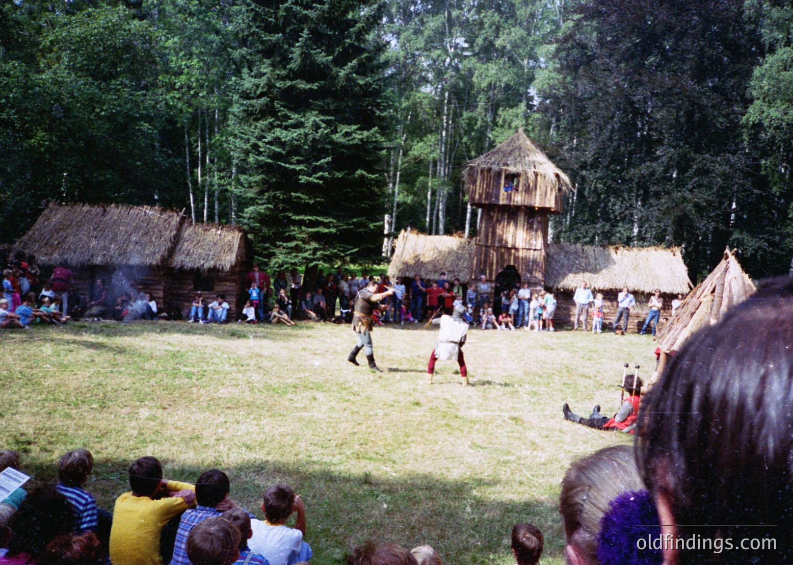 Traditional thatched huts and wooden structures in a forested setting, likely a cultural festival or reenactment. Central figures in period costumes perform a ritual or dance, surrounded by seated spectators. Mid-20th century European folk heritage scene.