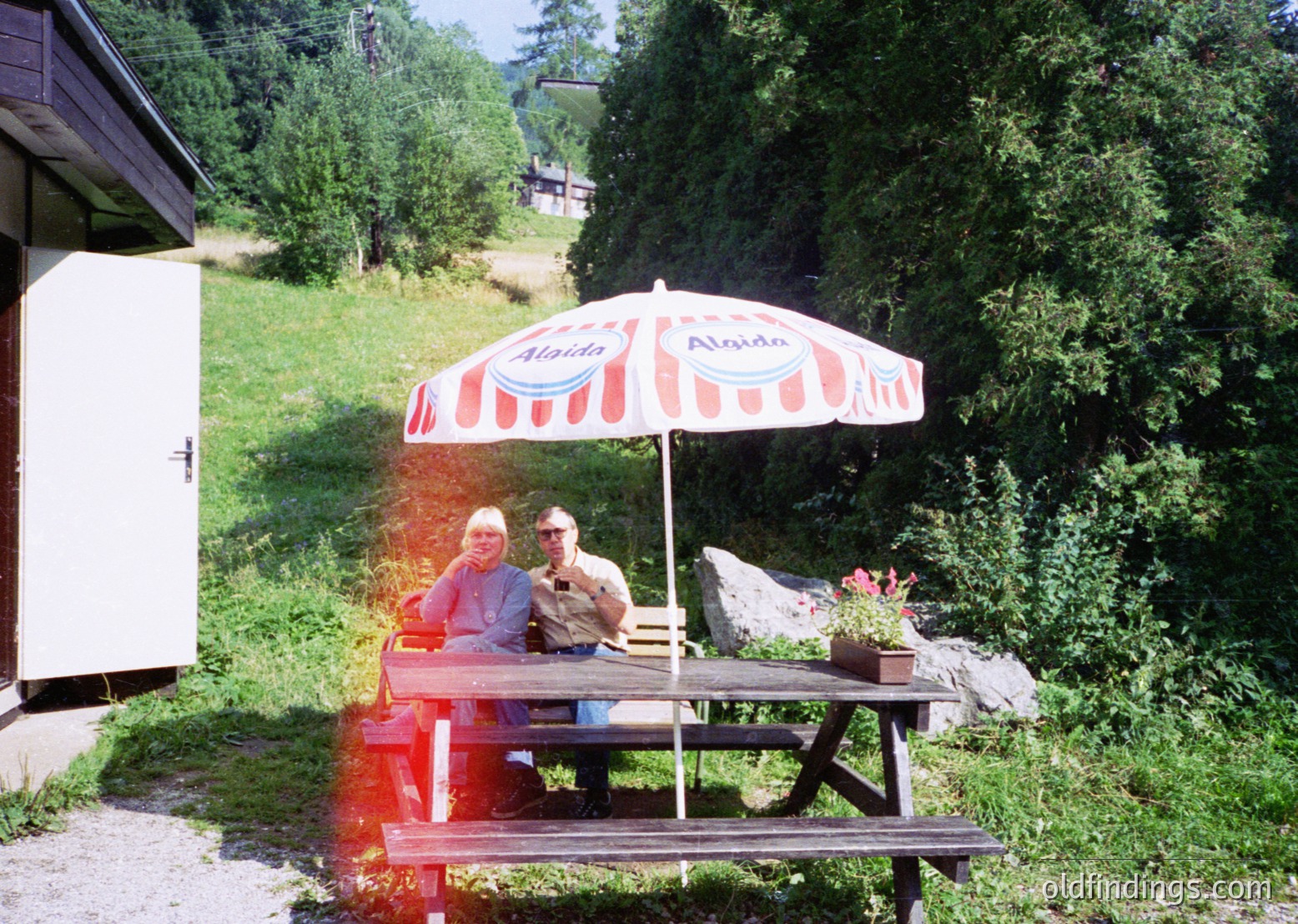 Two individuals relax under a vintage **Alpina** red-and-white striped umbrella at a rustic picnic table in a lush alpine setting. The scene captures mid-20th century outdoor leisure, likely the 1960s-70s, with wooden cabins and dense greenery in the background. Perfect for vintage travel, nostalgia, or outdoor lifestyle content.