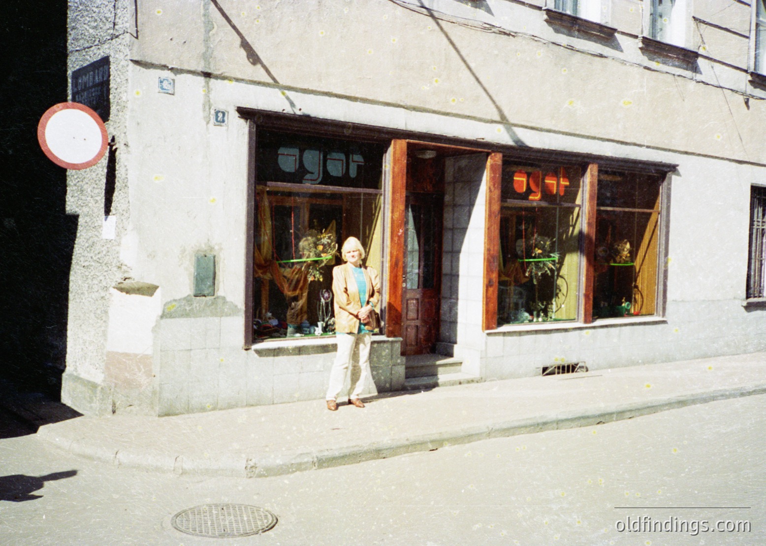 Retro storefront with Cyrillic sign "АГОРА" (Agora) in a European urban setting, likely 1970s-80s. Woman in light-colored blouse and trousers poses at entrance, surrounded by potted plants and vintage display items. Textured stone facade and street sign indicate historical architecture.
