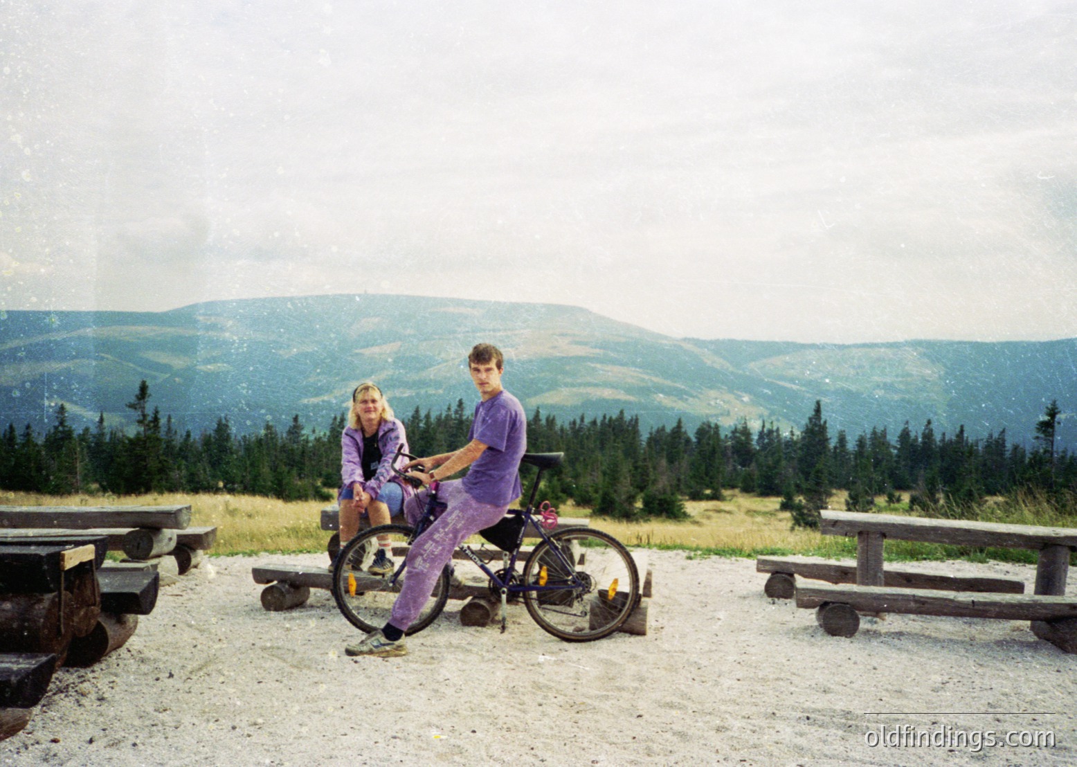 Two individuals pose on a gravelled overlook with mountain views, wearing matching purple cycling attire. The man holds a vintage-style bike with a basket. Wooden picnic tables and forested slopes frame the scene, suggesting a recreational area. Likely late 20th century, possibly or .