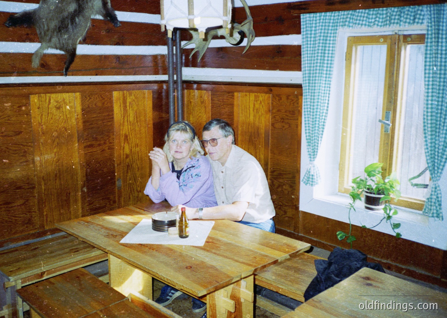 Two individuals pose indoors in a rustic wooden cabin, likely late 20th century. The man wears glasses and a white shirt; the woman wears a purple top. A mounted deer head and vintage tools hang above them. A small potted plant sits by the window, framed with green checkered curtains.