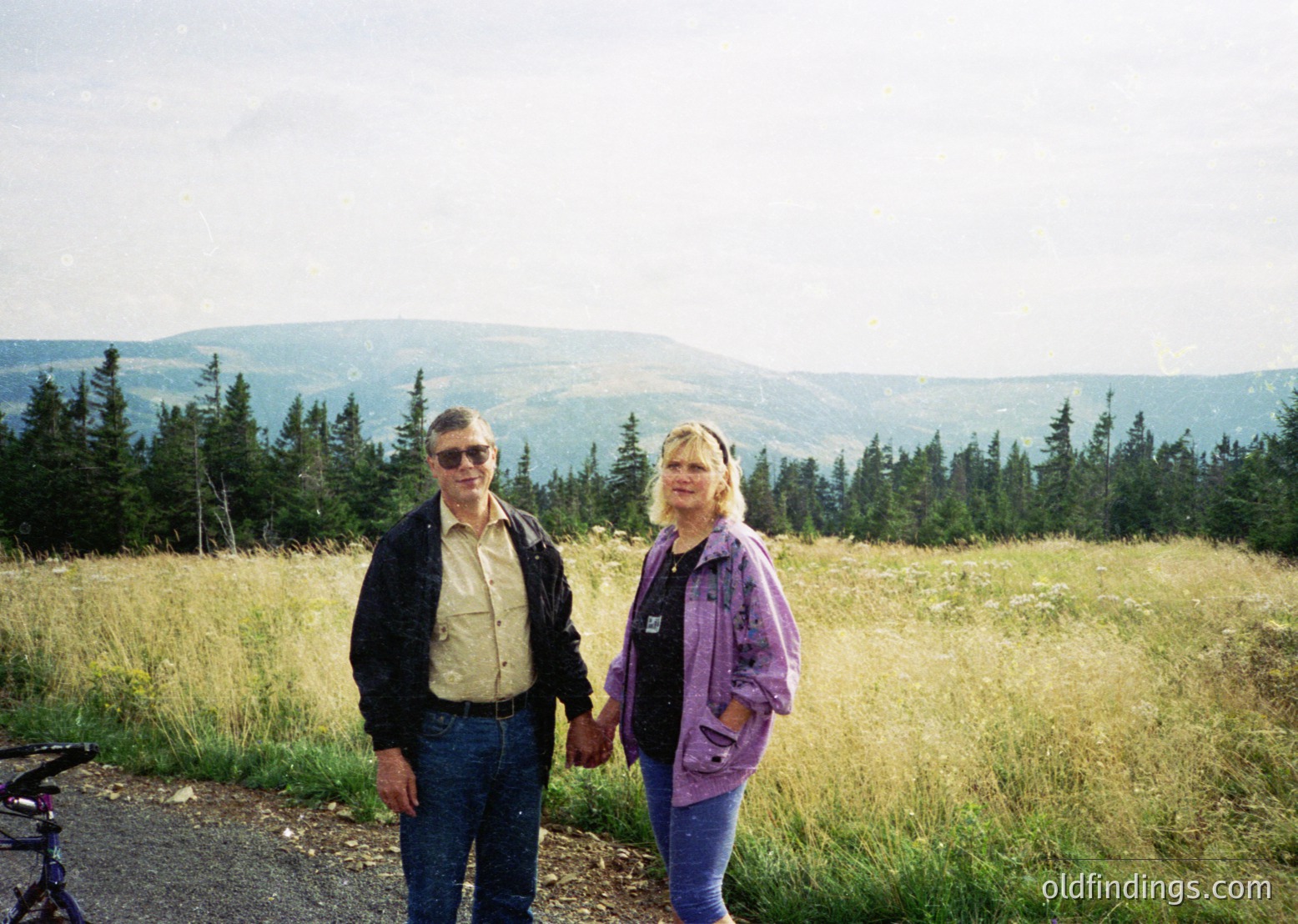 Couple posing on a scenic mountain road surrounded by dense coniferous forest and rolling hills. Man in sunglasses, dark jacket, and jeans; woman in purple jacket, light hair. Mid-20th century outdoor attire suggests or .
