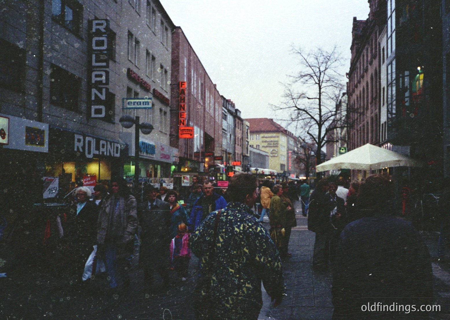 Vintage urban street scene with heavy snowfall, bustling pedestrians in winter attire. Prominent Roland and Pannen stores on brick-front buildings, likely European. Snow-covered sidewalks and leafless trees suggest late 20th-century city life.