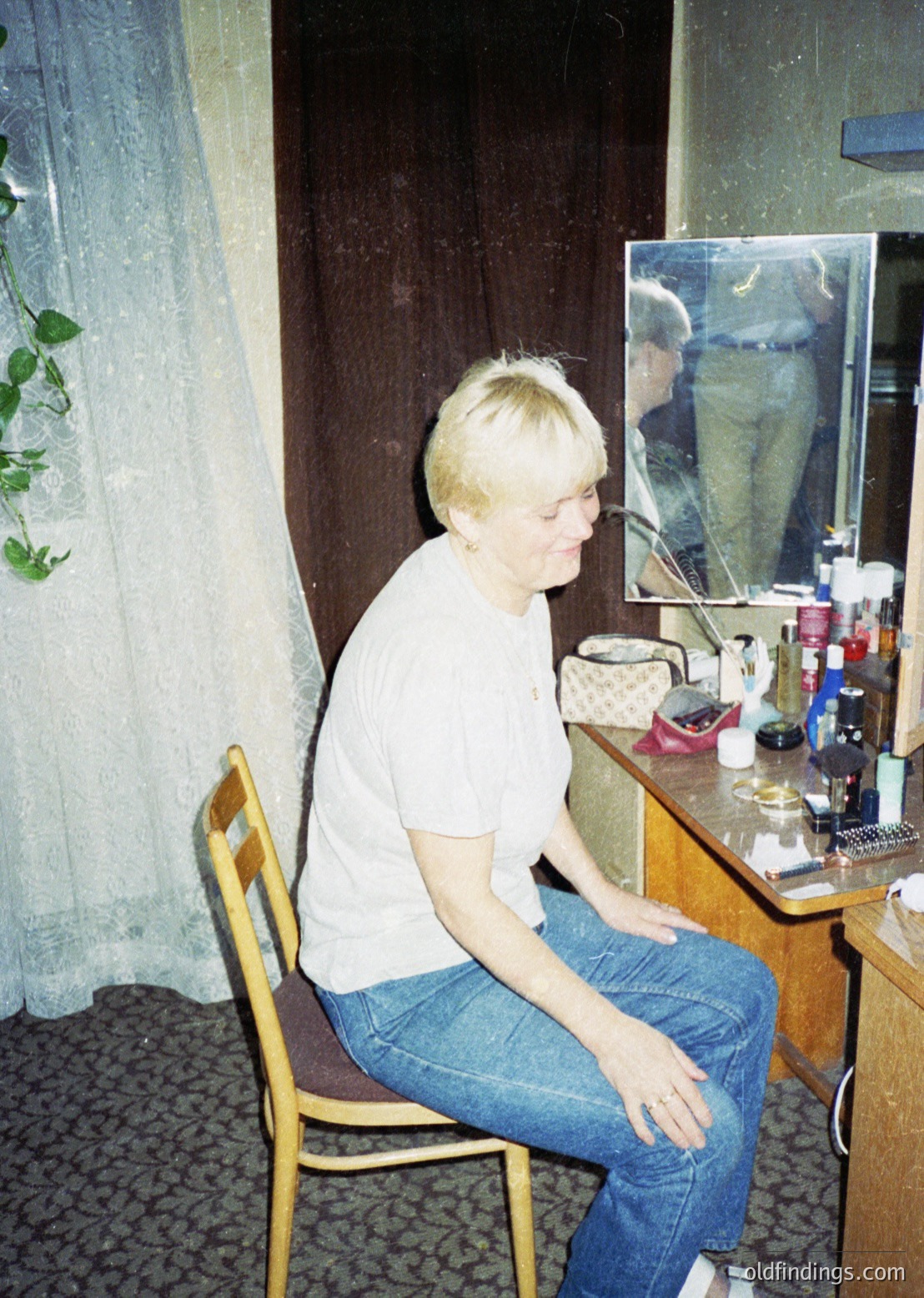 A person sits on a wooden chair in a vintage bathroom, adjusting their hair in front of a mirror with a small framed photo. Wooden vanity holds skincare bottles, a comb, and a small basket. Curtains with floral patterns frame the window.
