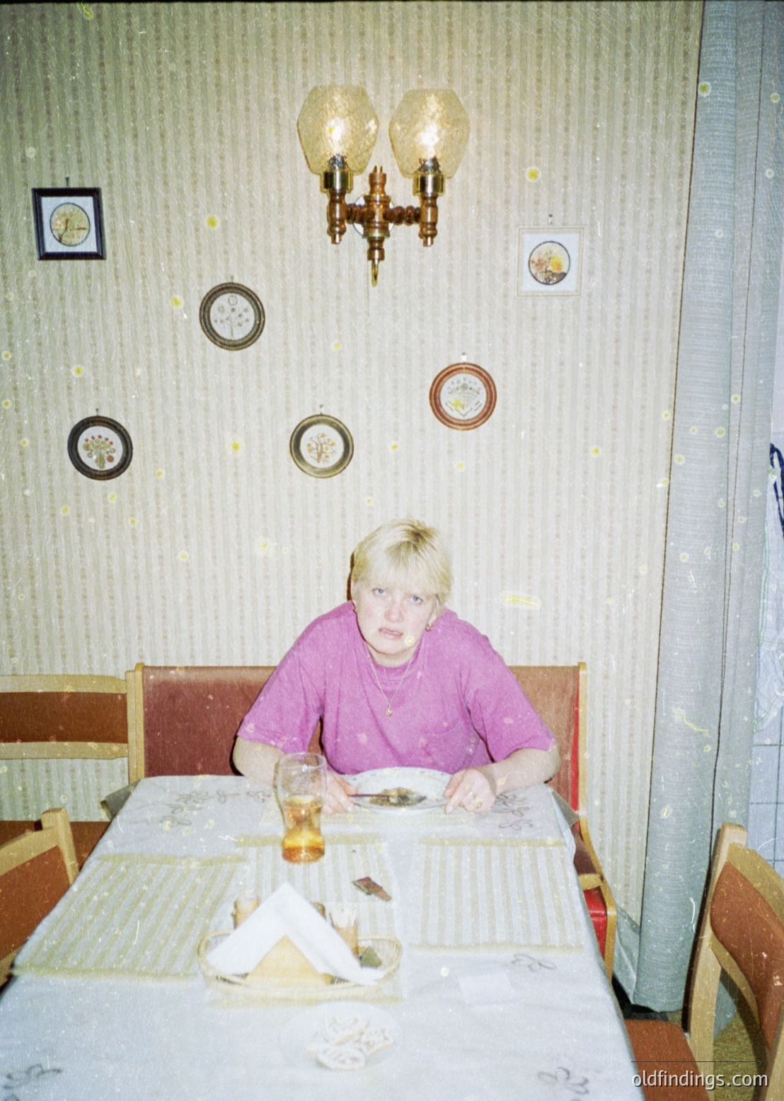 Vintage interior shot of a woman seated at a table in a retro diner-style setting. Decor includes a brass chandelier, round wall clocks, and patterned wallpaper. Table features a patterned tablecloth, glass, and a half-eaten meal.