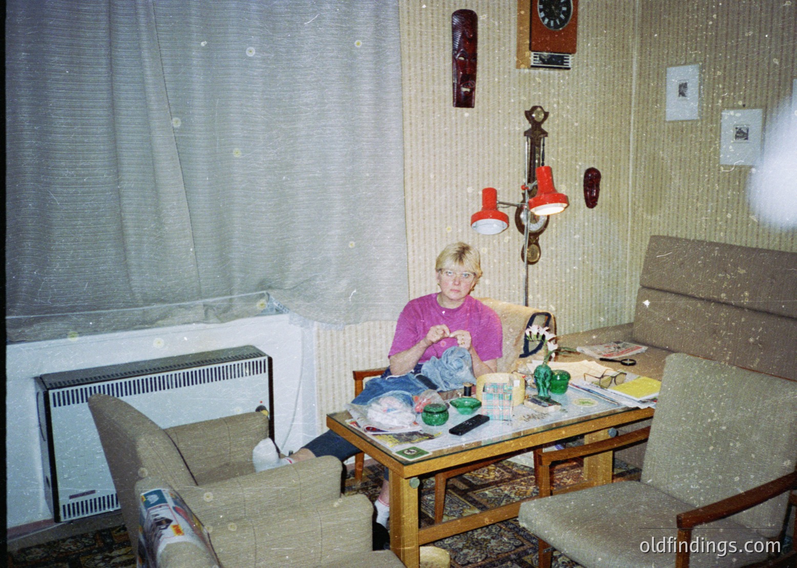 Vintage indoor scene featuring a child seated at a cluttered wooden table, surrounded by toys and household items. Mid-century design elements include a wall-mounted radiator, vintage lamp with red shade, and patterned sofa. Decor includes framed photos and a wall clock. Likely Eastern European residential interior, 1970s-1980s.