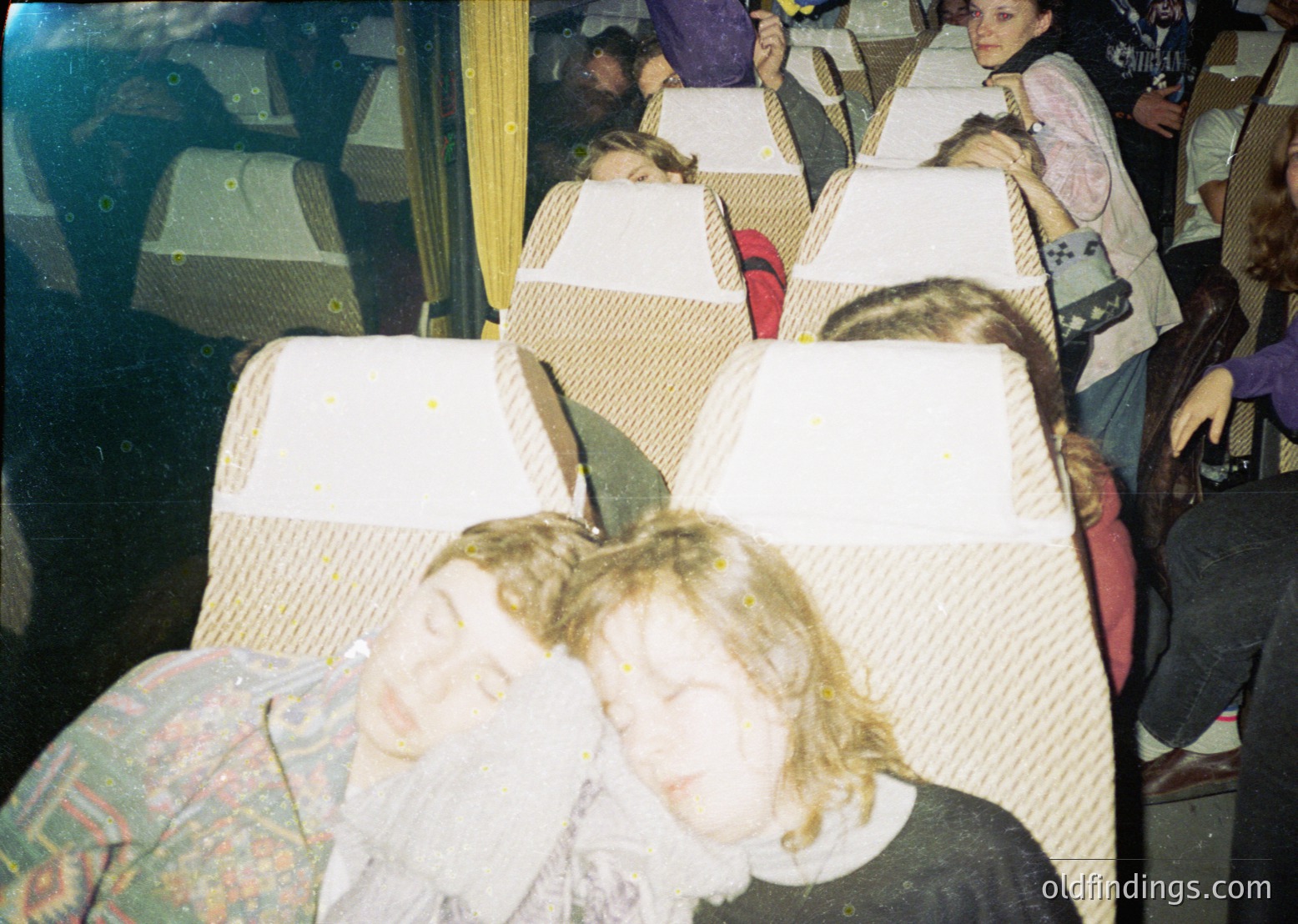 Vintage bus interior with passengers seated in rows of padded, beige upholstered seats. Blurred faces suggest motion or low-resolution film. Men’s patterned jackets and women’s headscarves hint at 1970s–1980s Eastern European style. Window reveals daylight, indicating daytime travel.