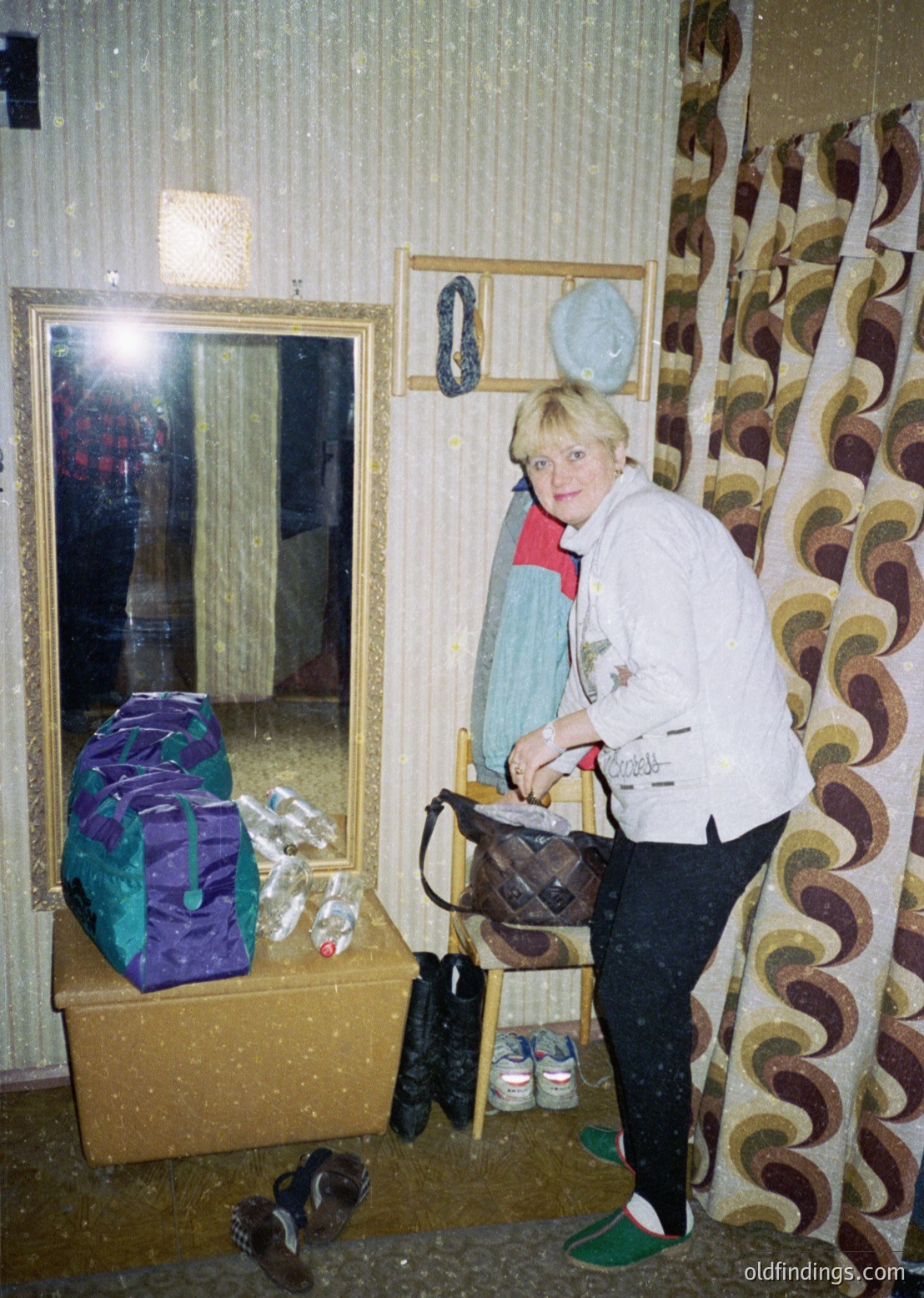 Woman in a retro-style sweater poses indoors beside a mirrored wall, holding a brown handbag. Surrounding her: a wrapped gift box, mismatched shoes, and a wall adorned with patterned wallpaper and circular decorative plates. Interior suggests mid-20th century European domestic setting.