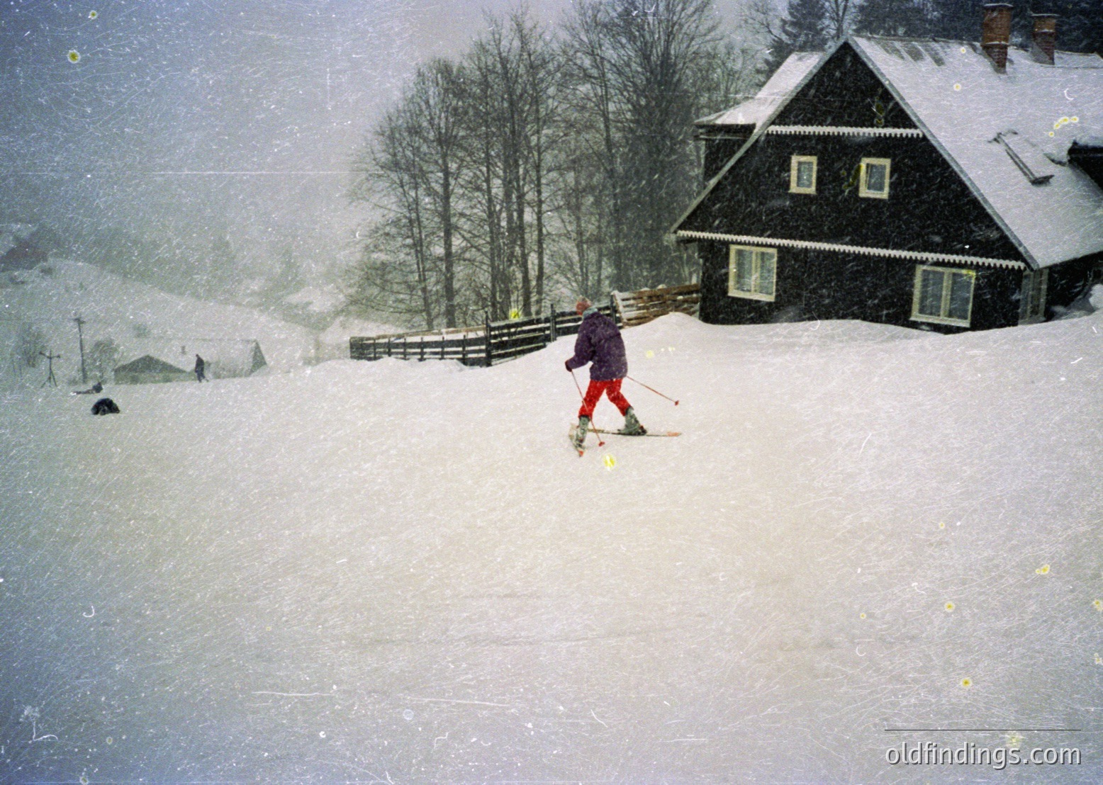 Vintage ski scene in heavy snowfall, featuring a lone skier in red pants and dark jacket navigating a groomed slope. Snow-laden wooden chalet with gabled roof and white-framed windows in background. Rustic wooden fence and forested hillsides enhance alpine atmosphere. Likely mid-20th century European ski resort.