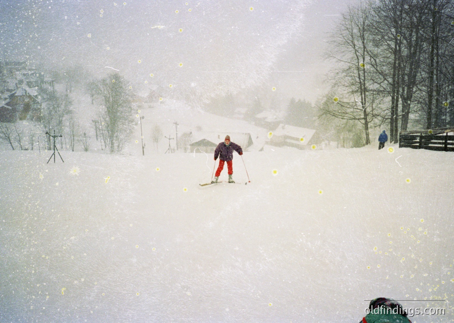 Snow-covered alpine slope with skier in red pants and dark jacket navigating heavy snowfall. Snow-laden trees and distant wooden structures suggest a rural ski resort. Blurred foreground indicates motion blur from camera or subject. Likely mid-20th century winter sports scene.