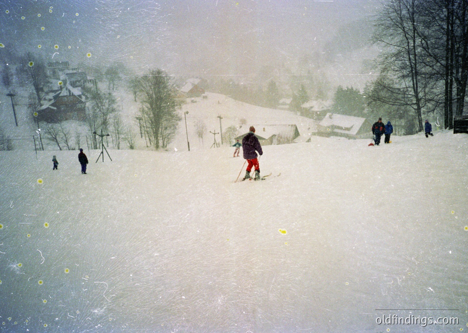 Vintage ski slope scene with heavy snowfall, featuring skiers in 1960s-70s gear. Snow-covered village and forested hills in background. Blurred motion captures dynamic winter activity.
