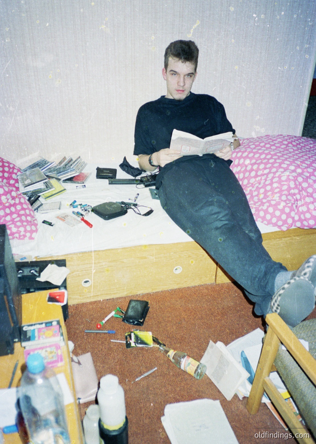 Young man reading in a cluttered, minimalist bedroom—1990s/early 2000s aesthetic. Notebooks, CDs, and a camera lie scattered on a wooden desk beside a single bed with floral bedding. Small bottles and tools hint at DIY or hobbyist interests.