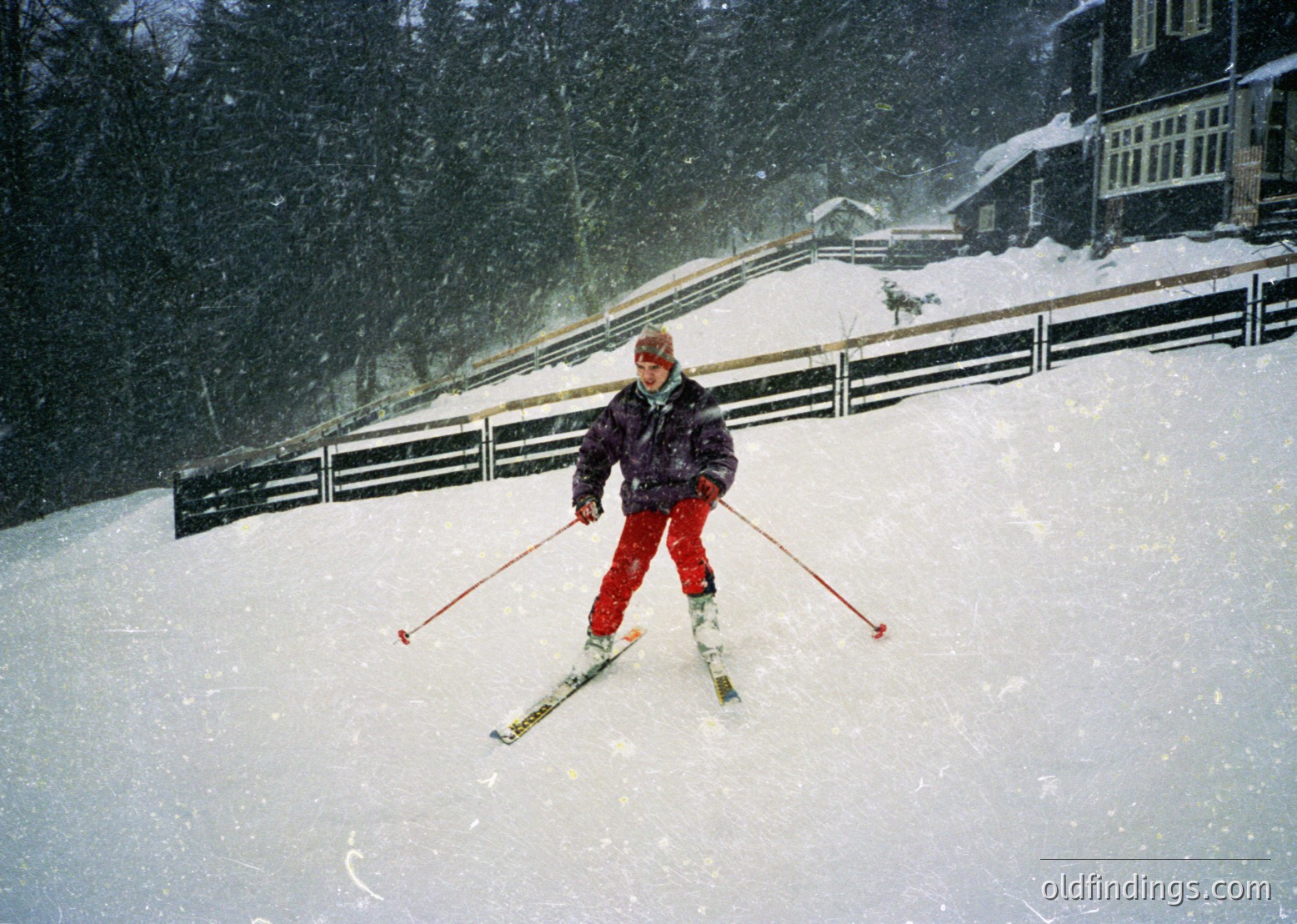 A skier in vintage alpine gear descends a snow-covered slope during heavy snowfall, framed by dense evergreen trees and a wooden fence. The mid-century ski attire and heavy snow suggest a or European alpine resort. Ideal for winter sports, vintage travel, and outdoor adventure content.