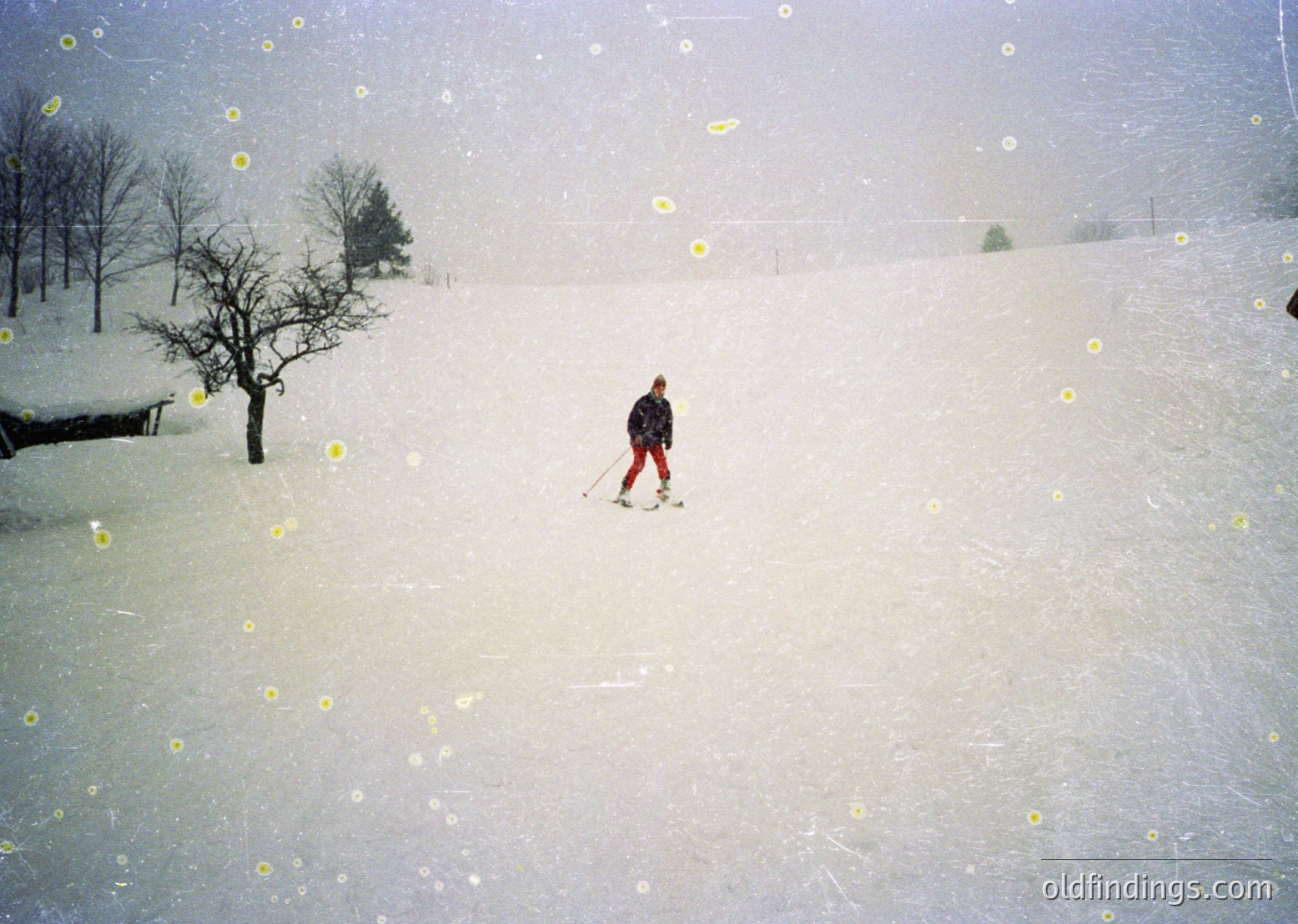 A lone skier in classic alpine gear descends a snow-covered slope during heavy snowfall, with sparse trees lining the background. The vintage filter suggests a mid-20th-century aesthetic, likely 1950s–1970s.