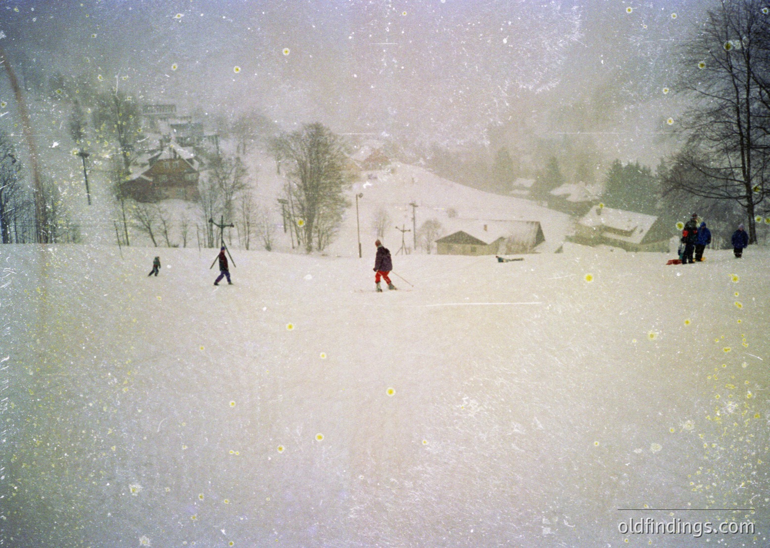 Winter scene featuring cross-country skiers navigating a snow-covered landscape. Snow-laden trees and rustic wooden buildings frame the scene, suggesting a rural alpine village. Heavy snowfall obscures distant details, emphasizing the serene yet dynamic atmosphere. Likely European alpine region, mid-20th century.
