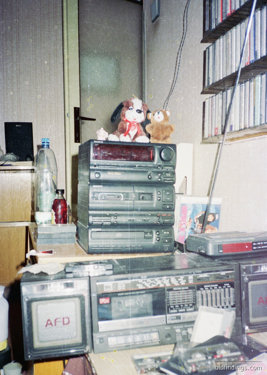 Vintage audio equipment stack with a plush dog perched atop, likely 1980s–1990s. Includes cassette decks, a boombox, and a CRT TV with "AFD" branding. Surrounding clutter suggests a retro electronics repair or hobbyist workspace.