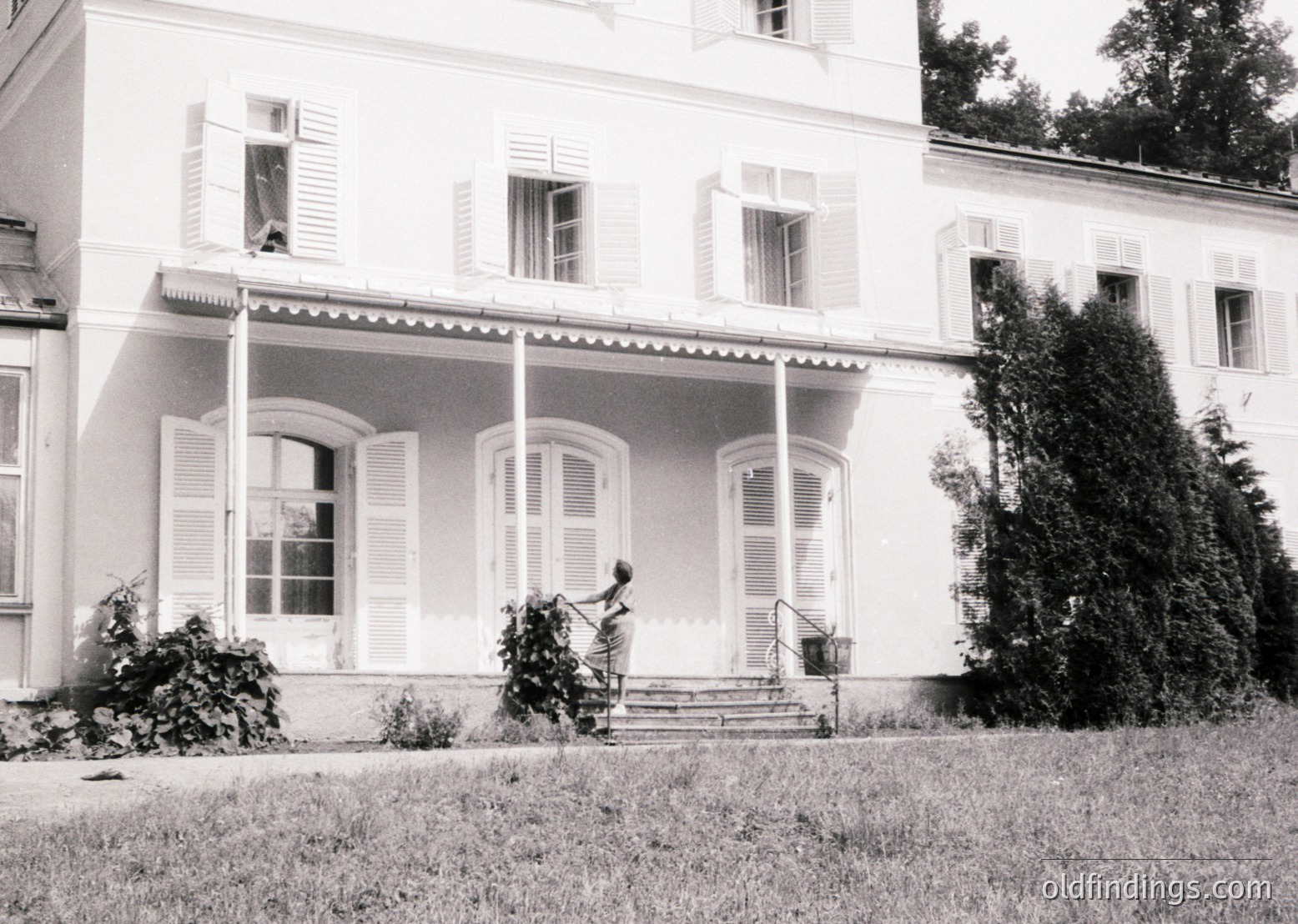 Neoclassical villa with symmetrical façade, shuttered windows, and a central staircase. A woman in period attire tends to potted plants near the entrance, framed by manicured greenery. Likely mid-20th century European residential architecture.
