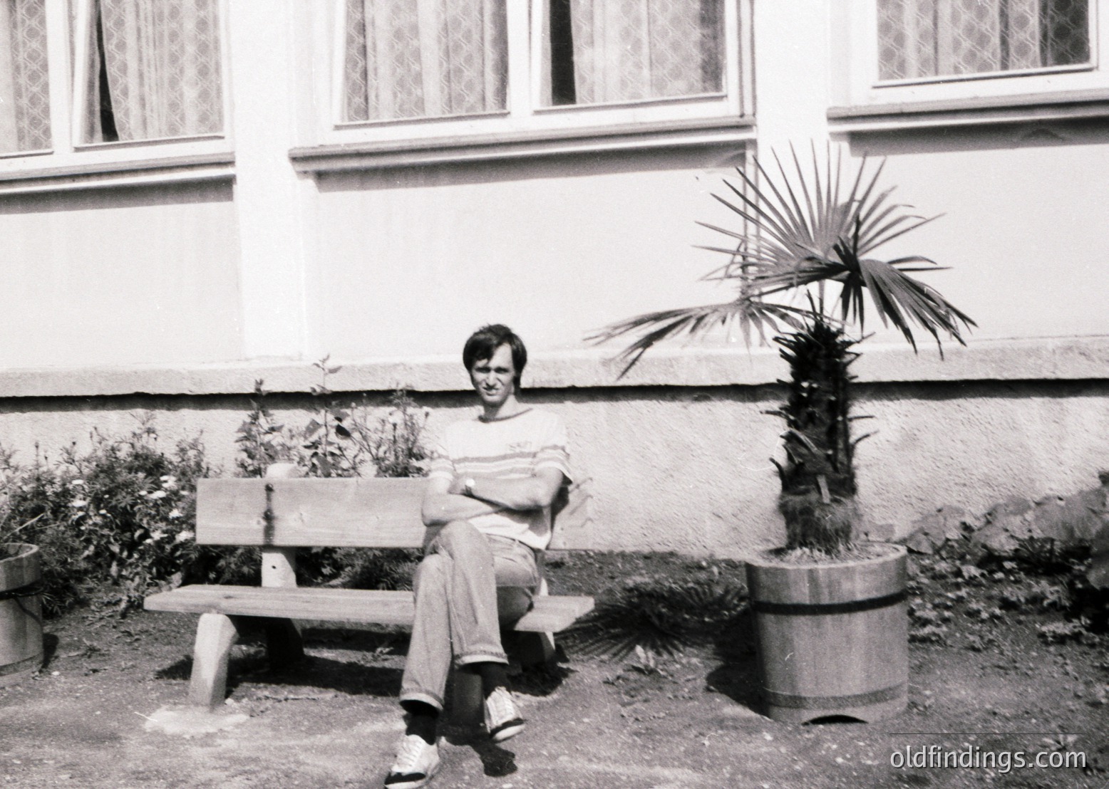 Black-and-white portrait of a person seated on a simple wooden bench in an urban courtyard. The individual wears a striped shirt, rolled-up trousers, and sneakers, holding a small object in their lap. A potted palm plant stands beside a wooden barrel planter. The building’s white facade features small, rectangular windows with curtains. Likely mid-20th century, possibly or .